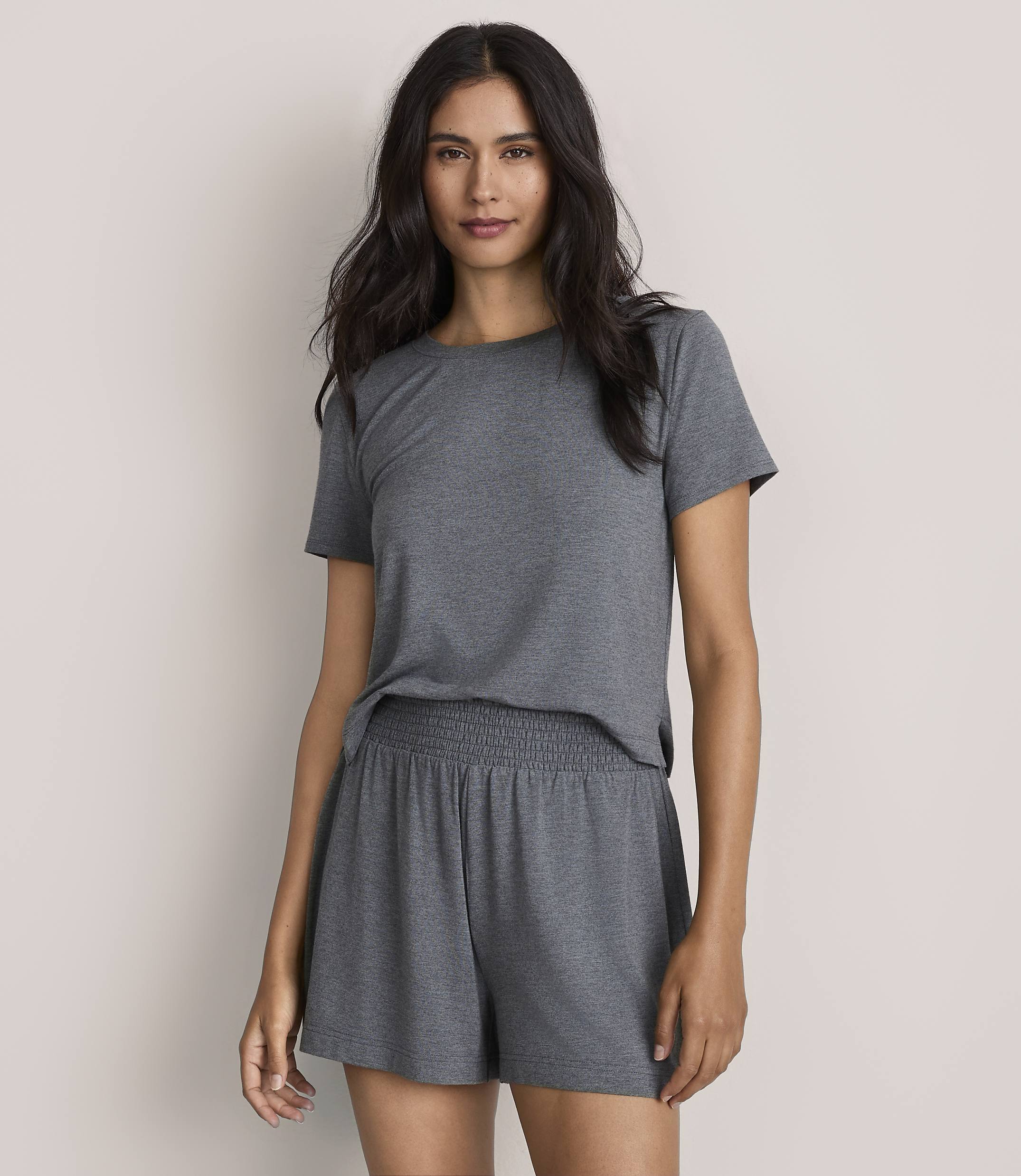 A woman wearing a grey t-shirt and matching shorts stands against a plain, light-colored background.