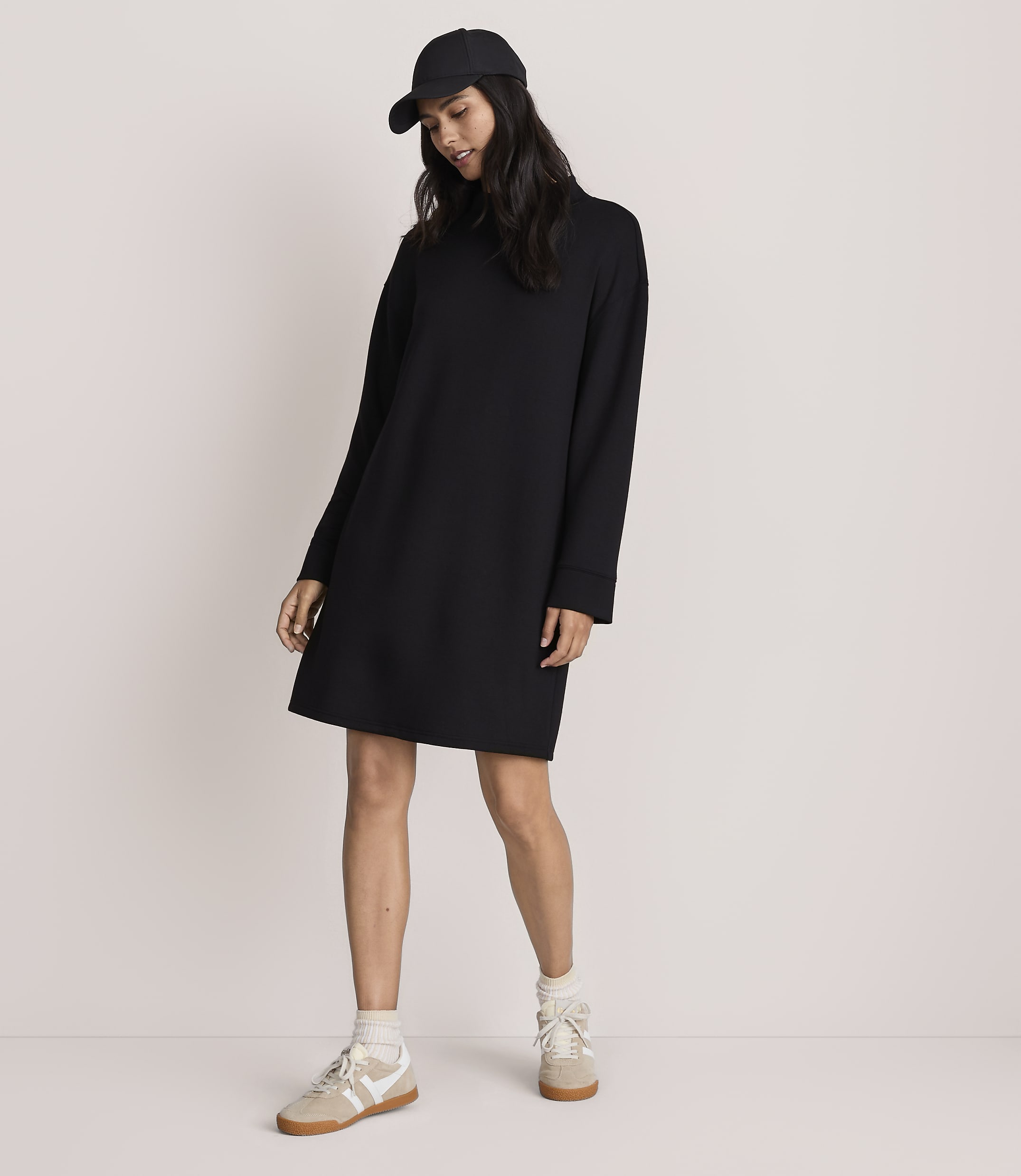 A woman wearing a black baseball cap, a long-sleeved black dress, and beige sneakers stands against a plain, light-colored background.
