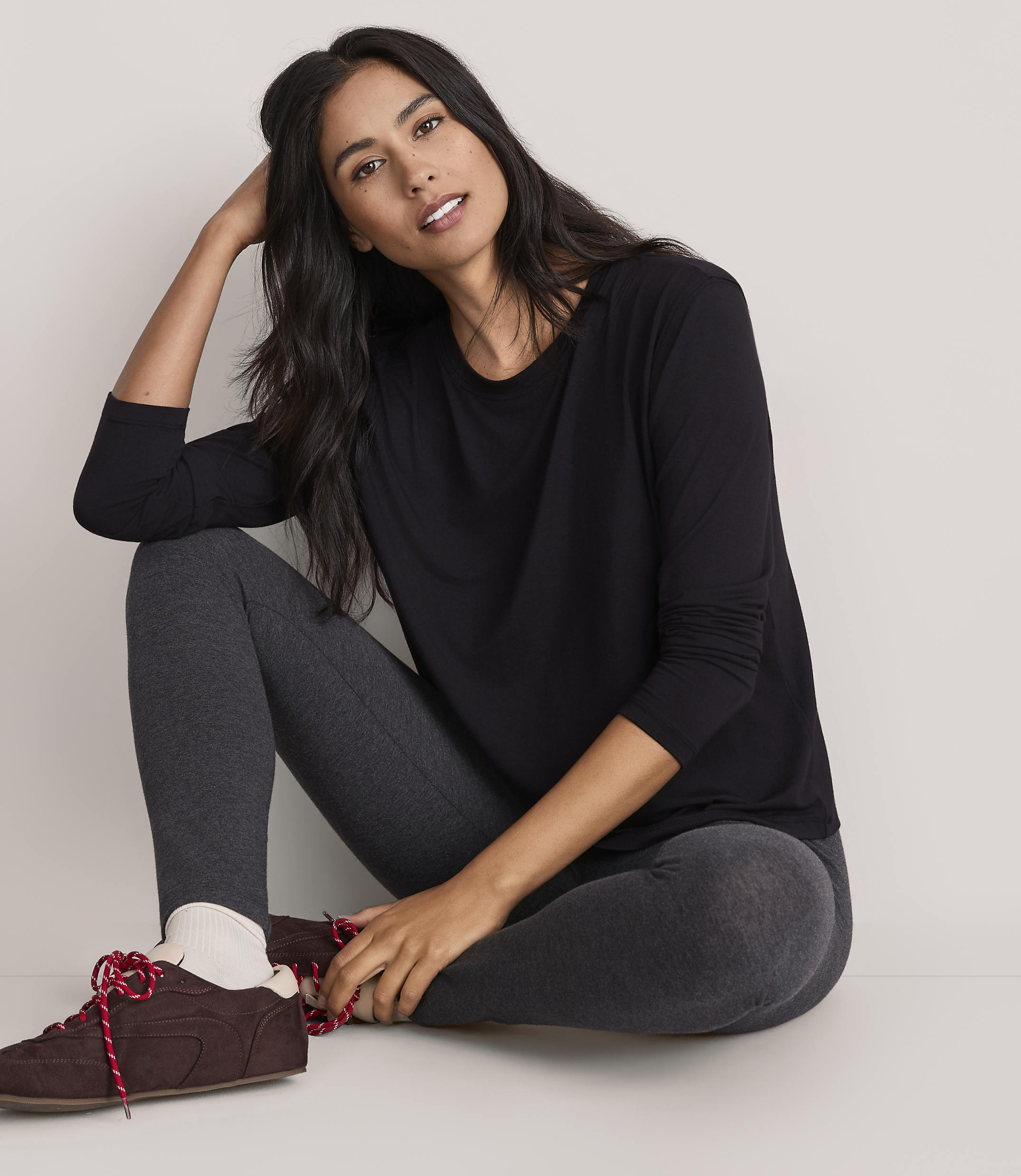 A woman wearing a black long-sleeved shirt and dark grey leggings sits on the floor against a plain, light-colored background.