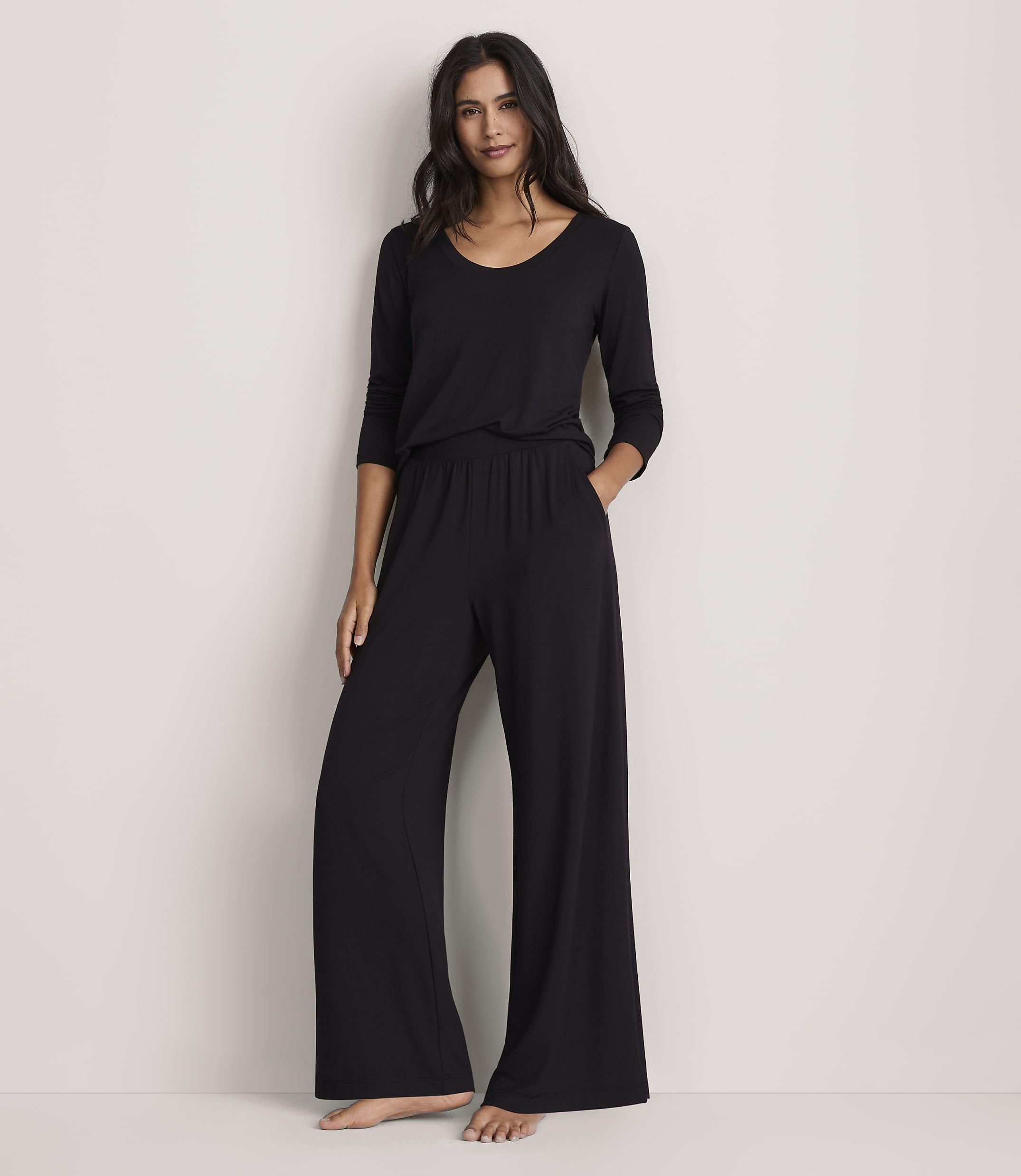 A woman wearing a black long-sleeved top and wide-leg black trousers stands against a plain, light-colored wall.
