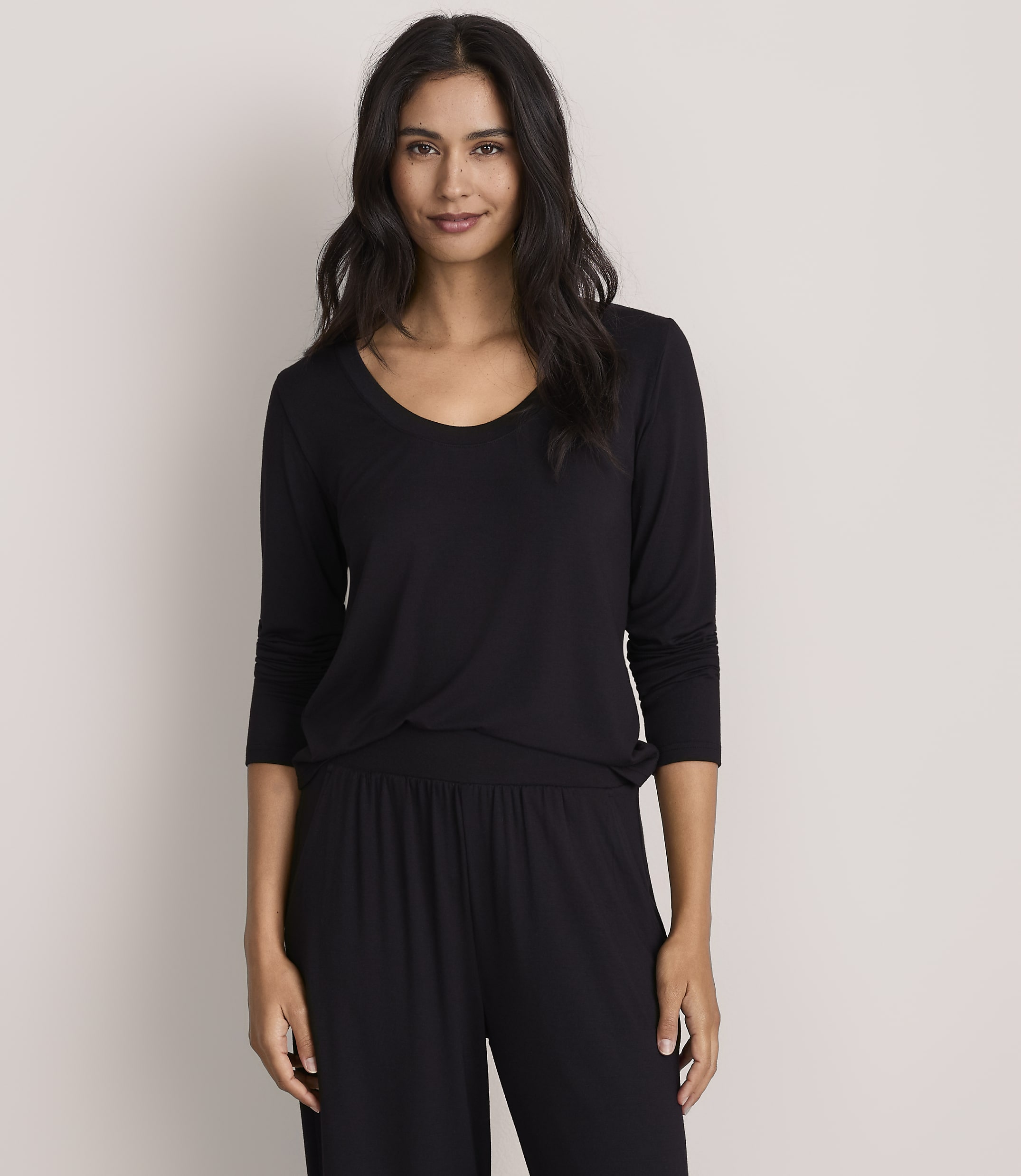 A woman with long dark hair wearing a black long-sleeved top and matching pants stands against a plain, light-colored background.