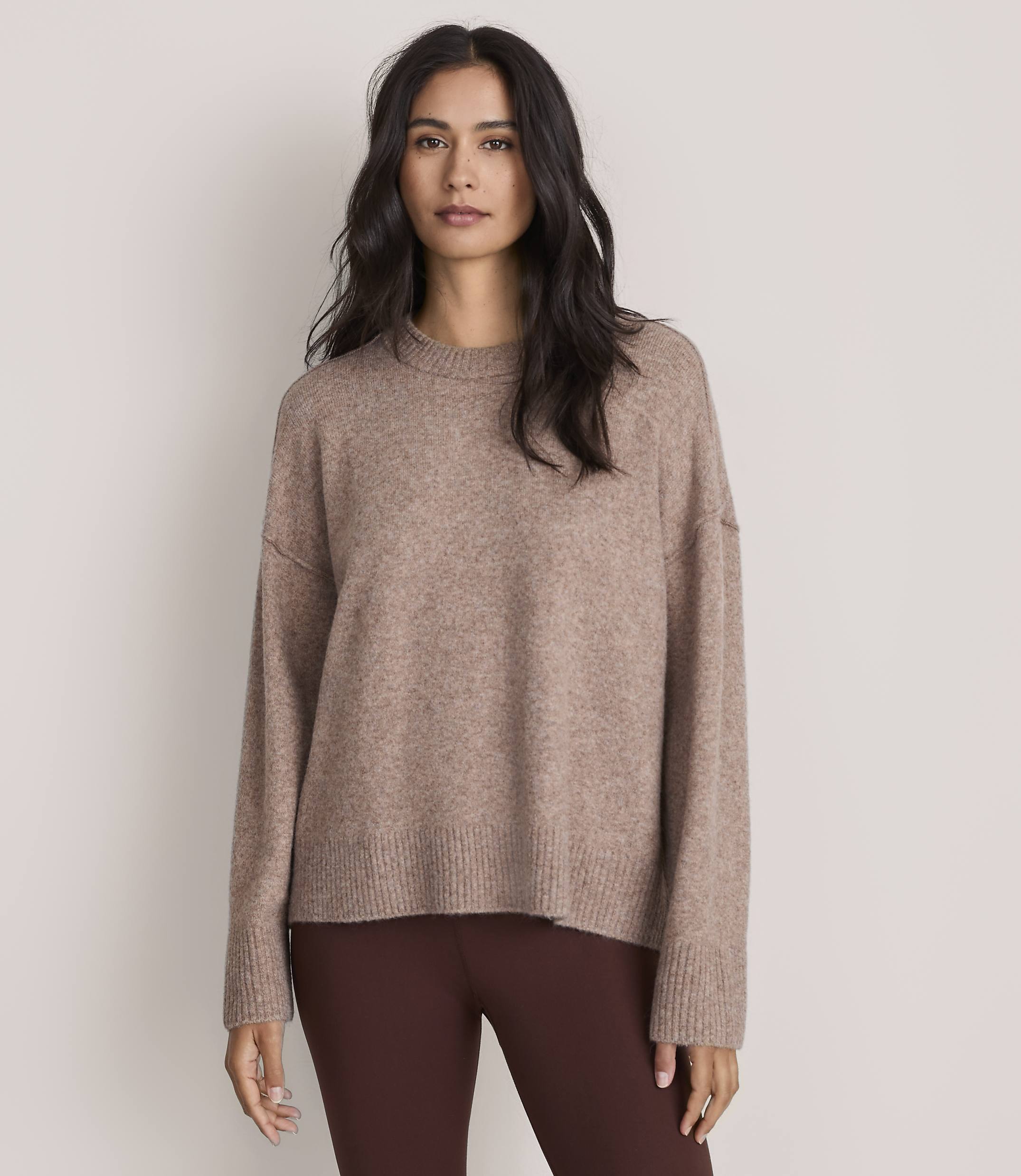 A woman wearing a brown oversized sweater and dark brown leggings stands against a plain, light-colored background.