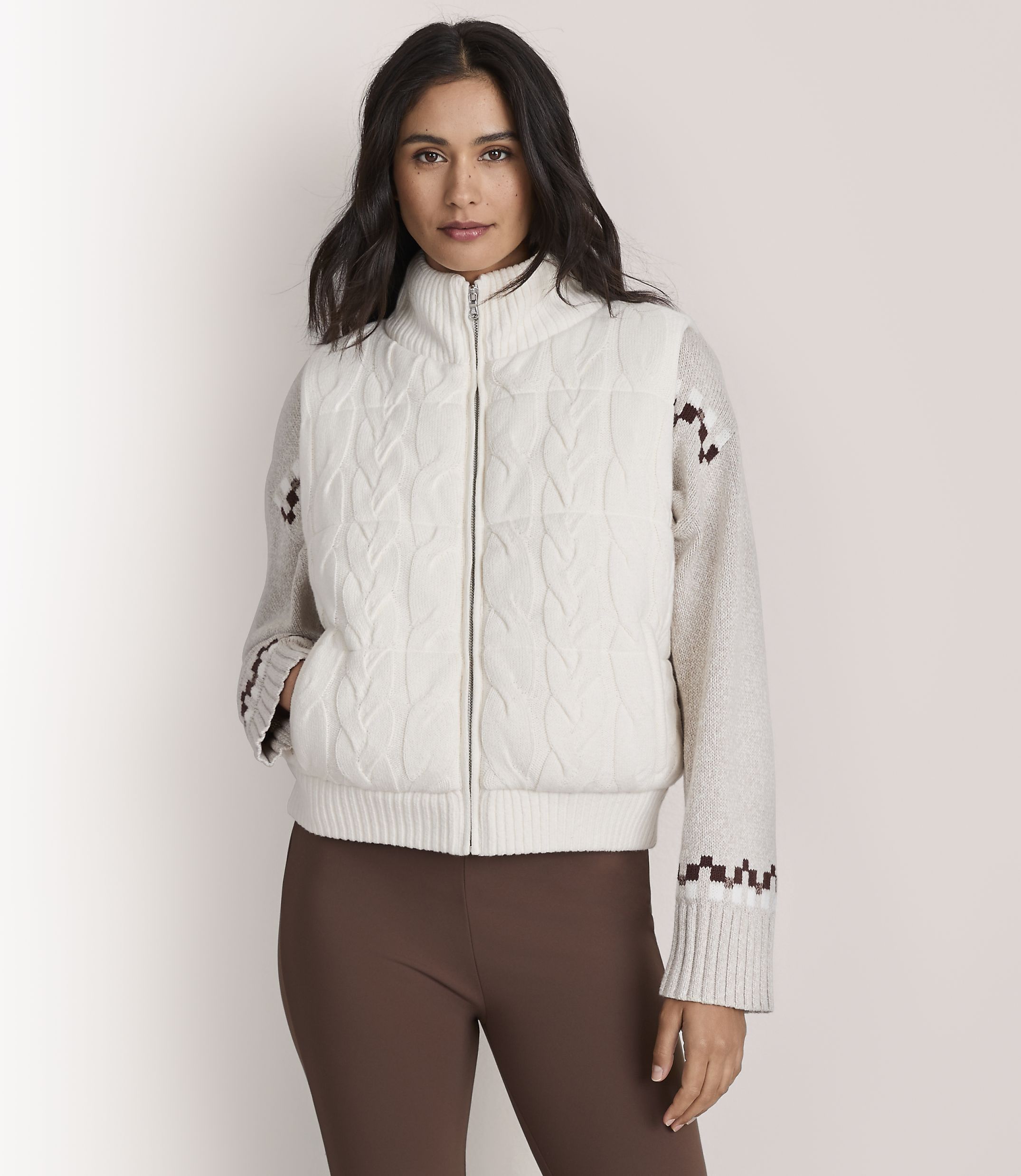 A woman wearing a white cable-knit zip-up sweater and brown leggings stands against a plain, light-colored background.