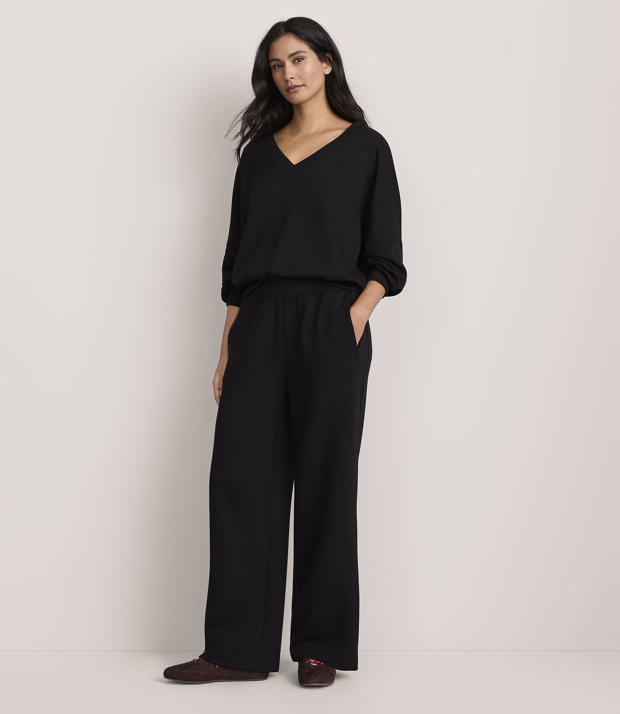 A woman wearing a black V-neck top and wide-leg black trousers stands against a plain, light-colored background.