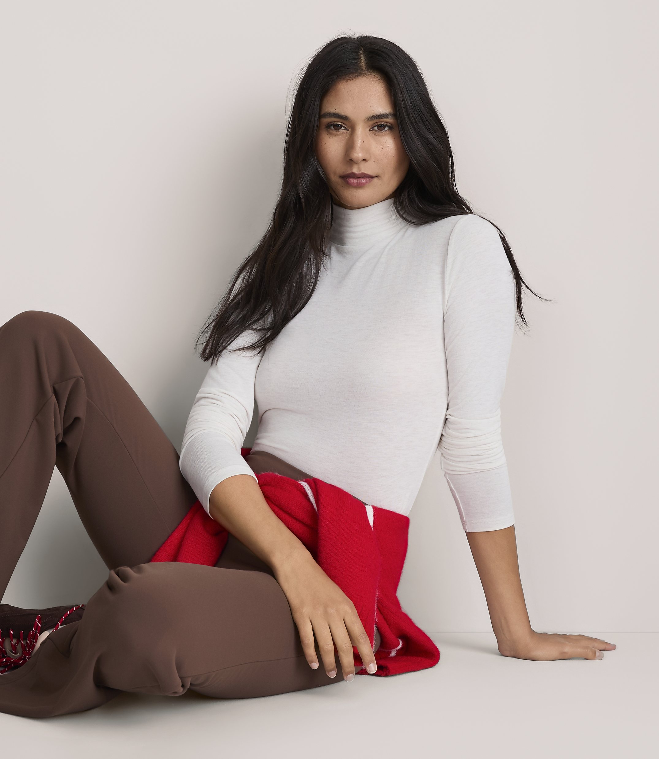 A woman wearing a white turtleneck and brown pants sits against a plain, light-colored background.