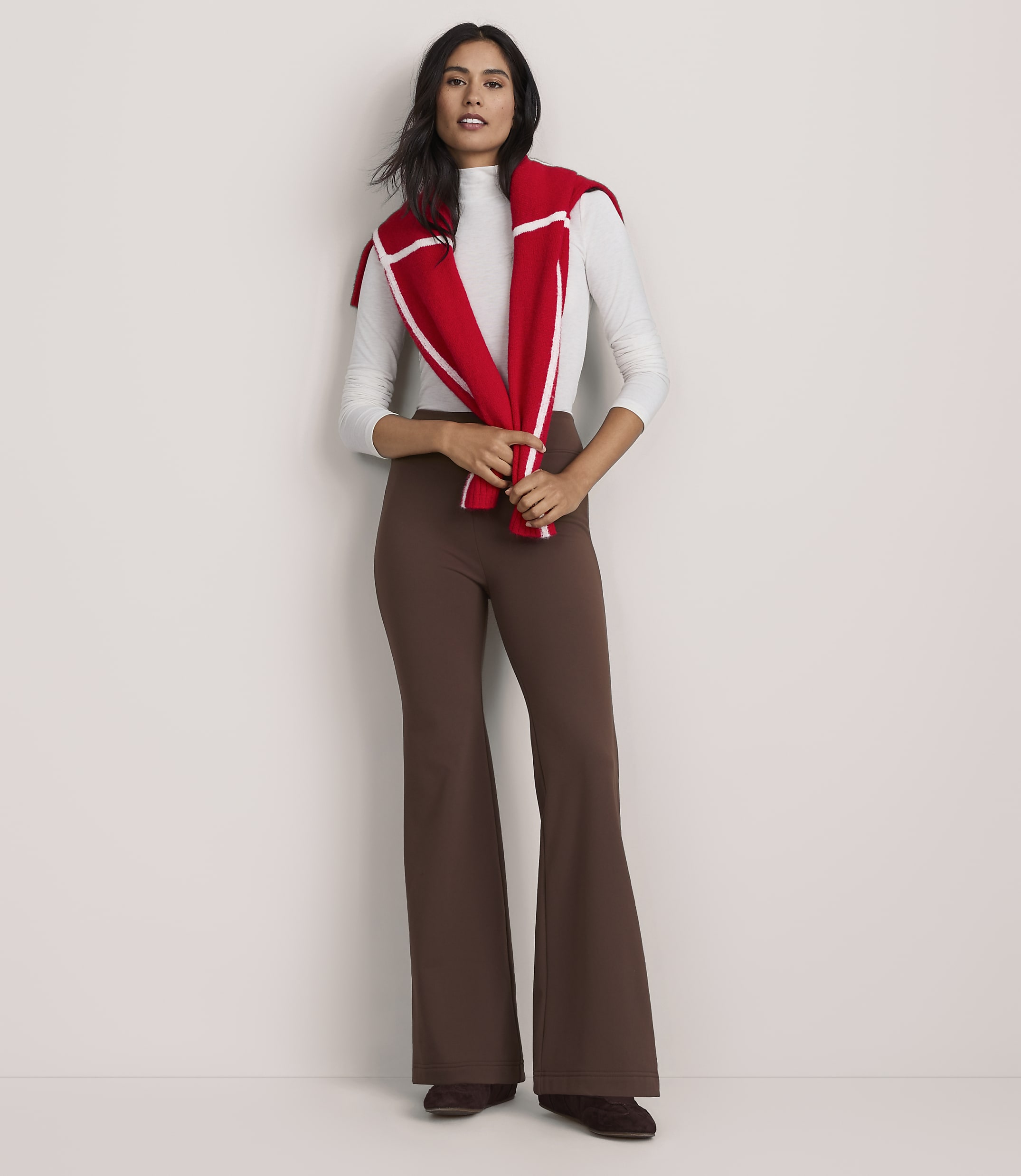 A woman wearing a white top, brown flared trousers, and a red sweater draped over her shoulders stands against a plain white background.