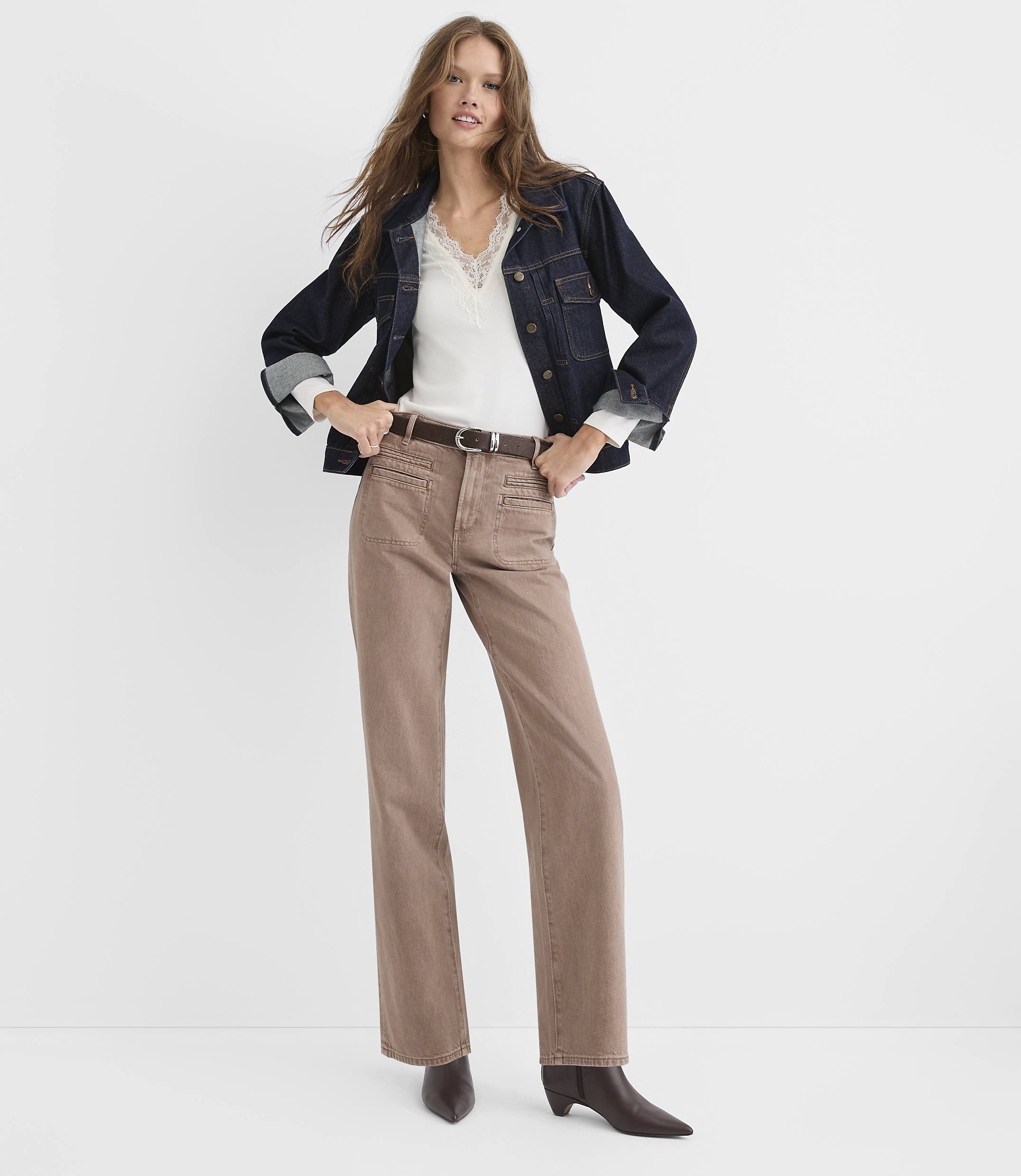 A woman wearing a dark denim jacket, white lace-trim top, and brown trousers stands against a plain white background.