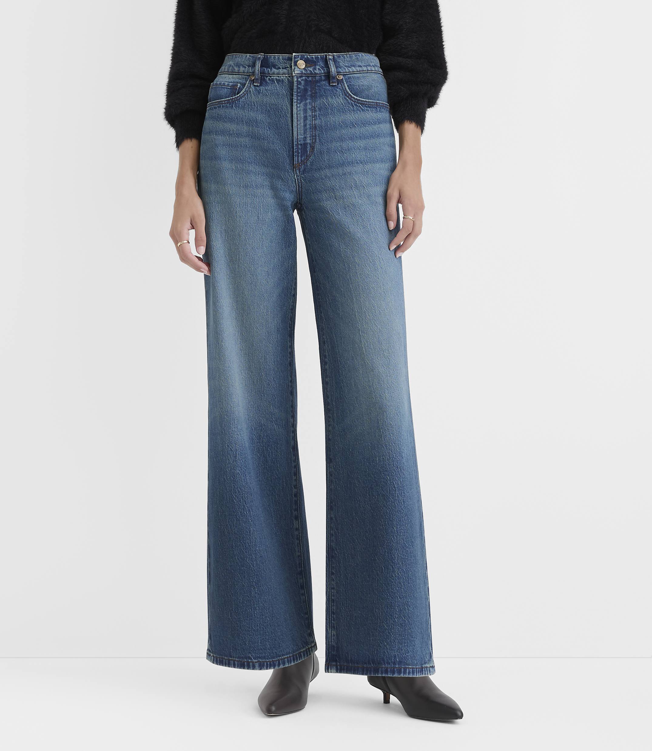 A person wearing a black sweater, wide-leg blue jeans, and black pointed-toe heels stands against a plain light gray background.