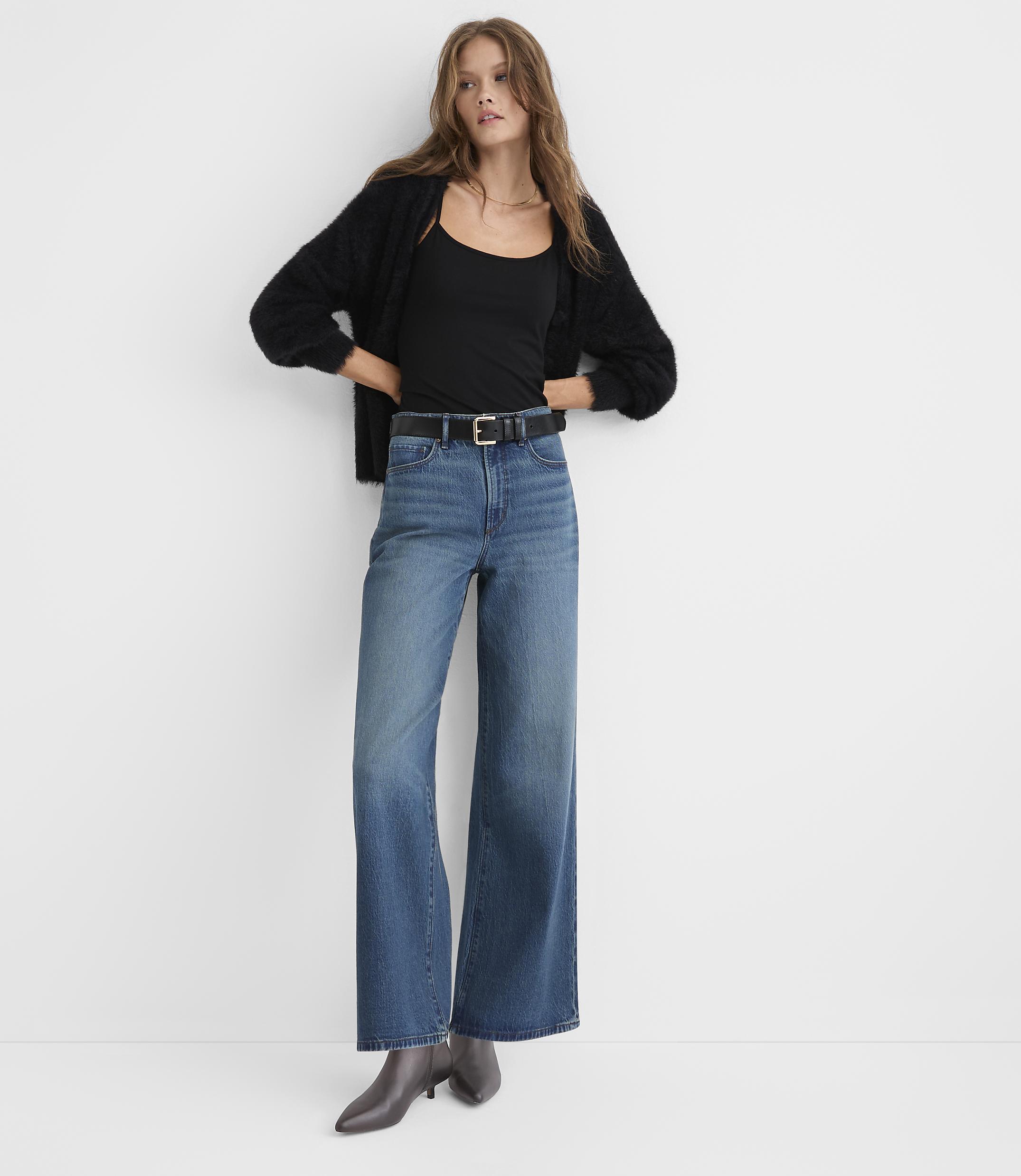 A woman wearing a black cardigan, black top, and wide-leg blue jeans stands against a plain white background.