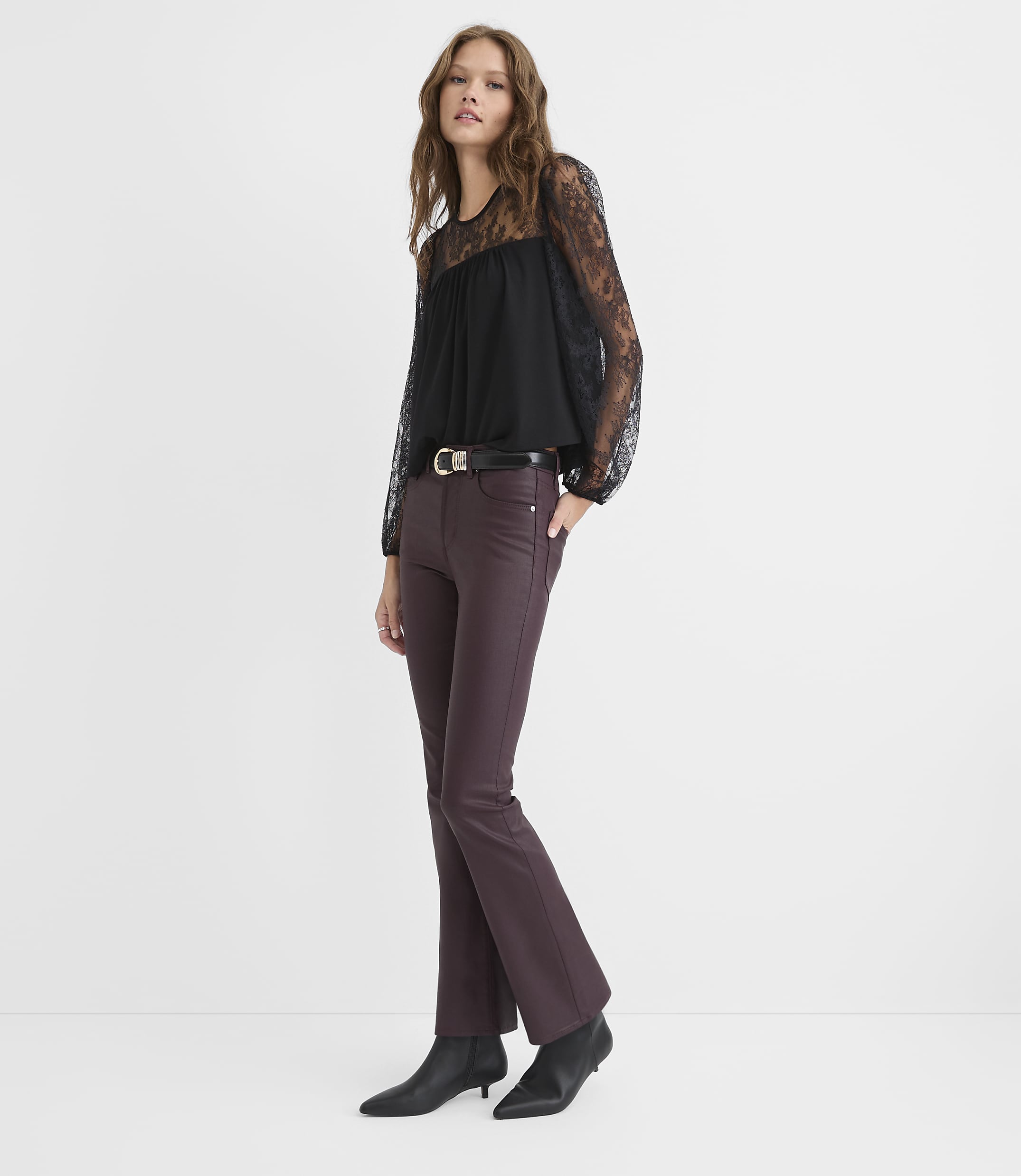A woman wearing a black lace-sleeved top and burgundy flared trousers stands against a plain white background.