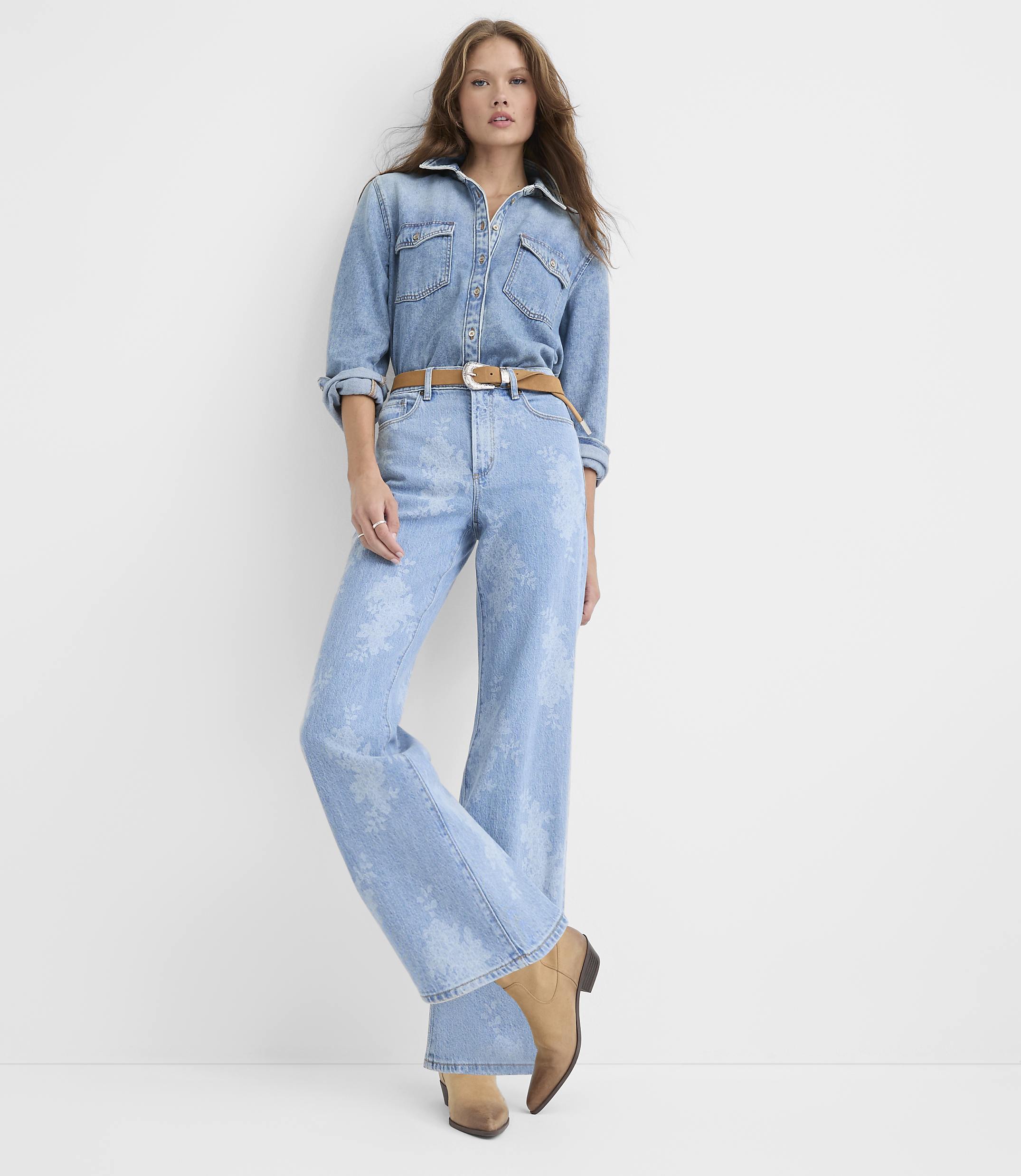 A woman wearing a light-wash denim shirt and matching wide-leg jeans with a brown belt stands against a plain white background.