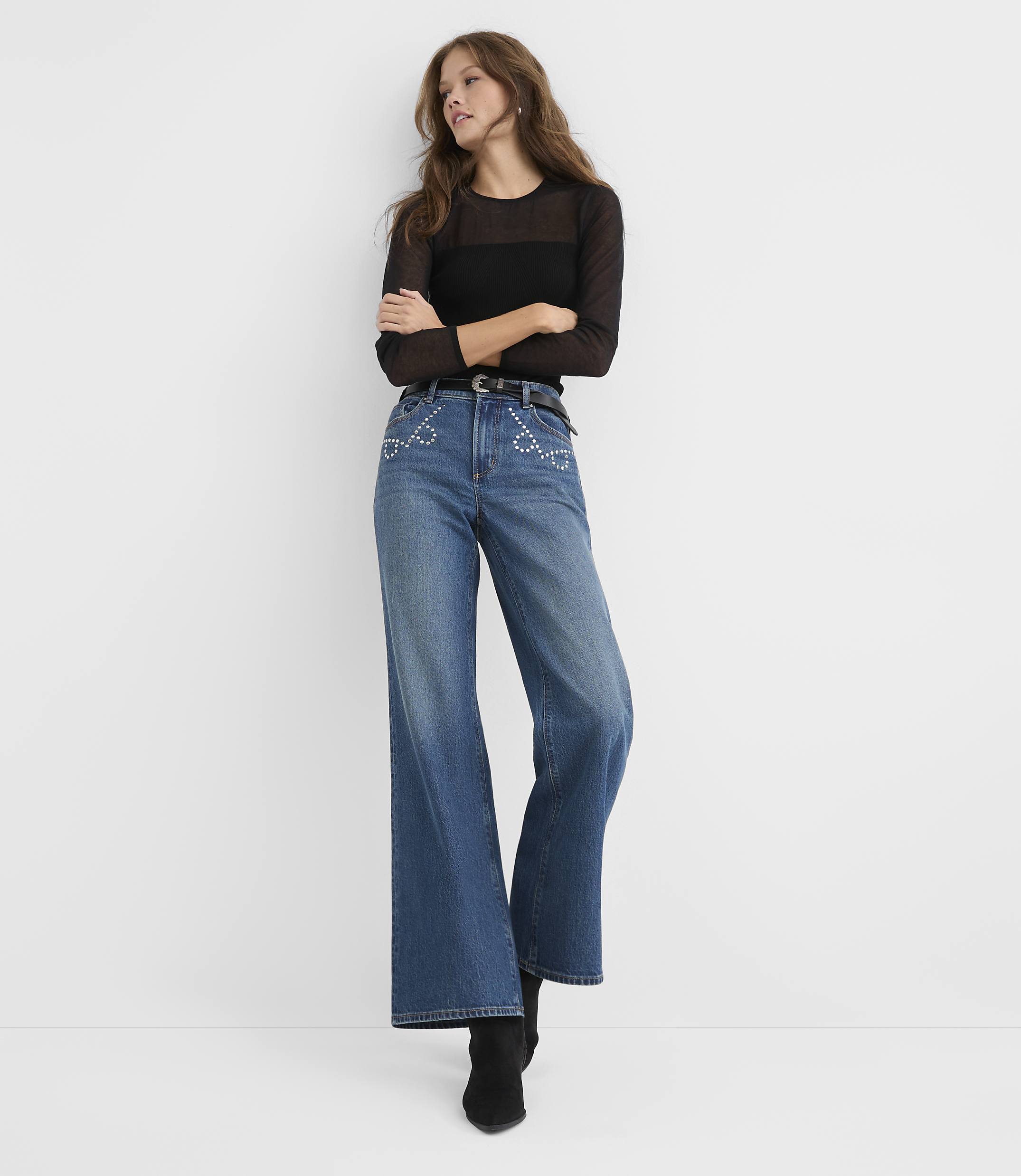 A woman wearing a sheer black top and wide-leg blue jeans stands against a plain white background.