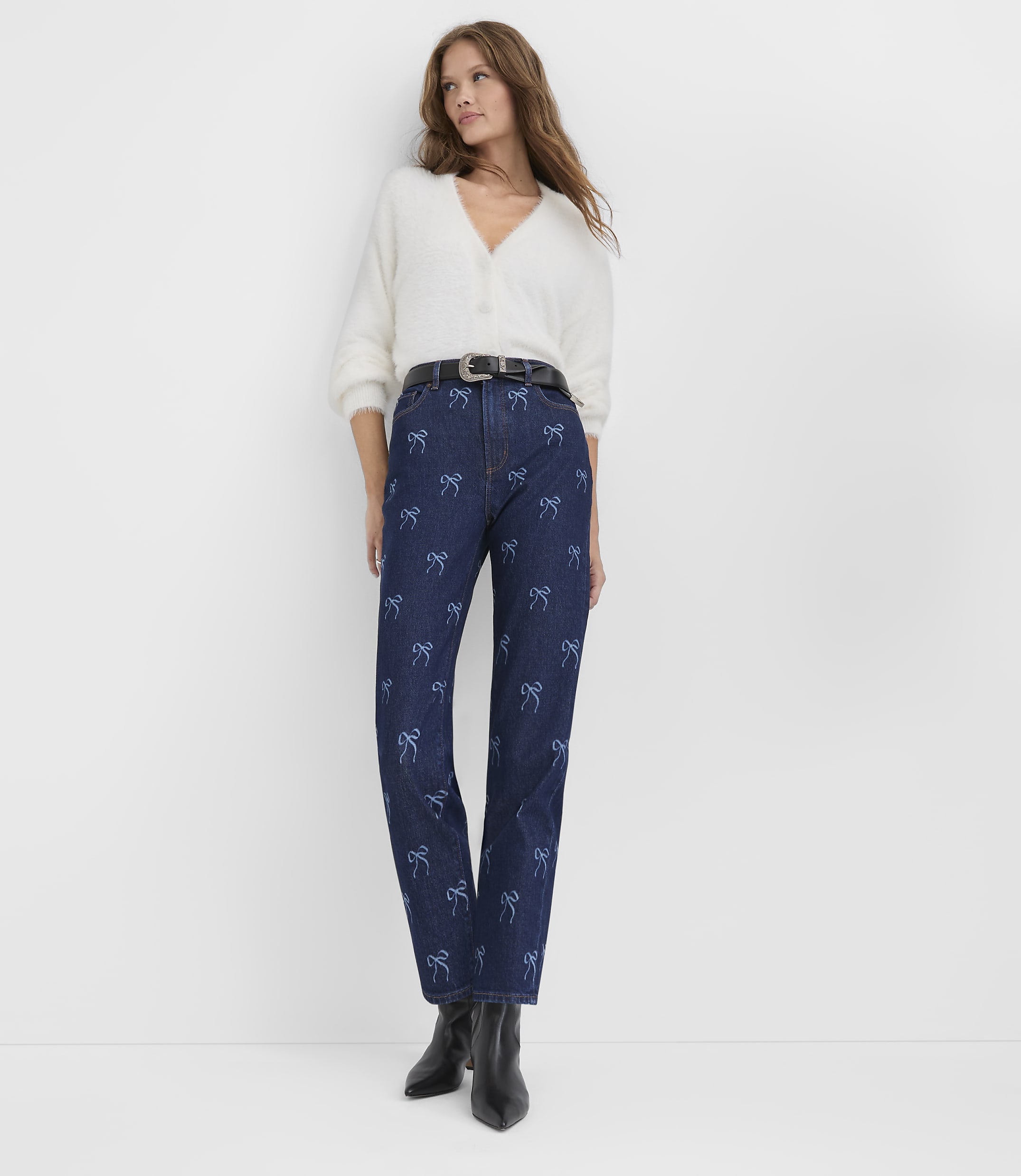 A woman wearing a white fuzzy cardigan and blue bow-patterned jeans stands against a plain white background.