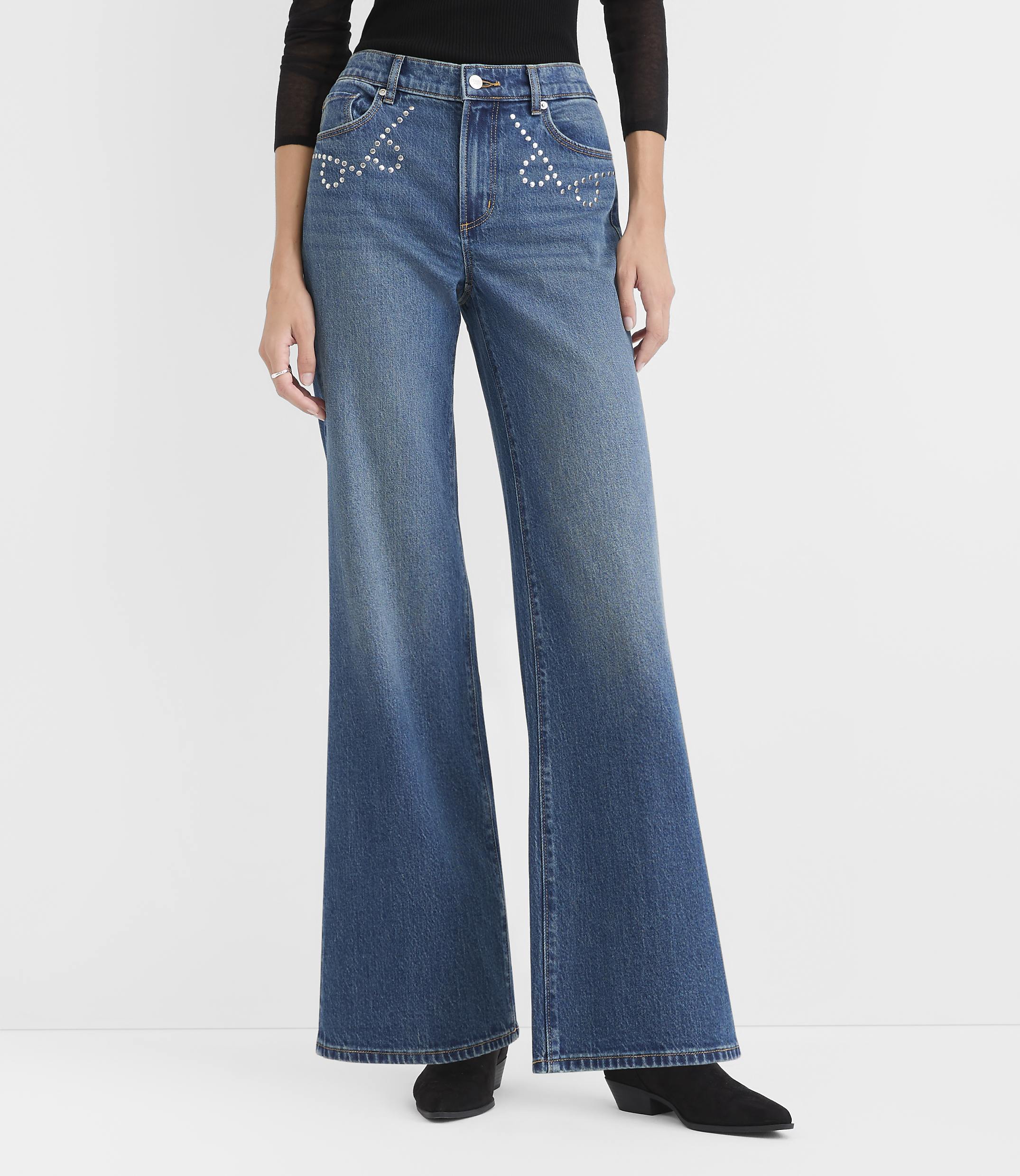 A person wearing a black top and wide-leg blue jeans with rhinestone heart details stands against a plain white background.