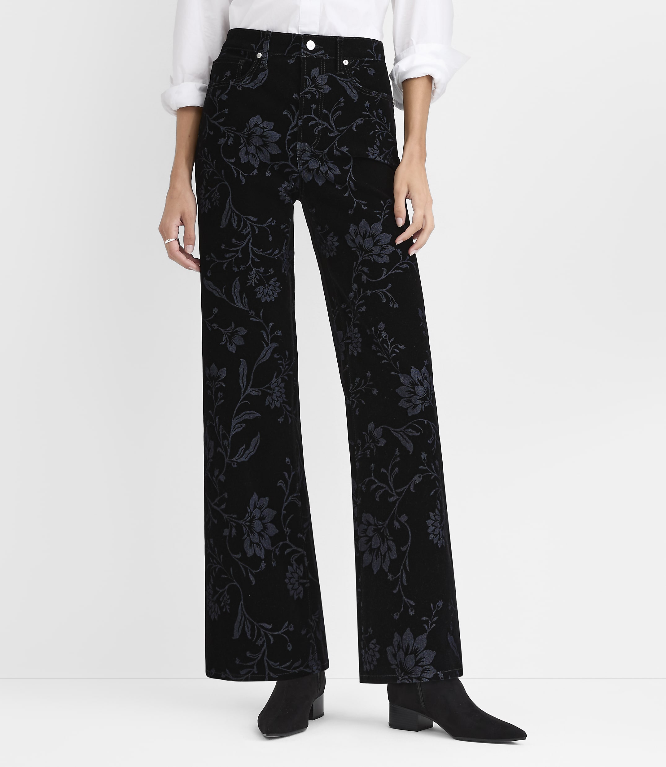 A person wearing a white shirt and black floral-patterned wide-leg pants stands against a plain white background.