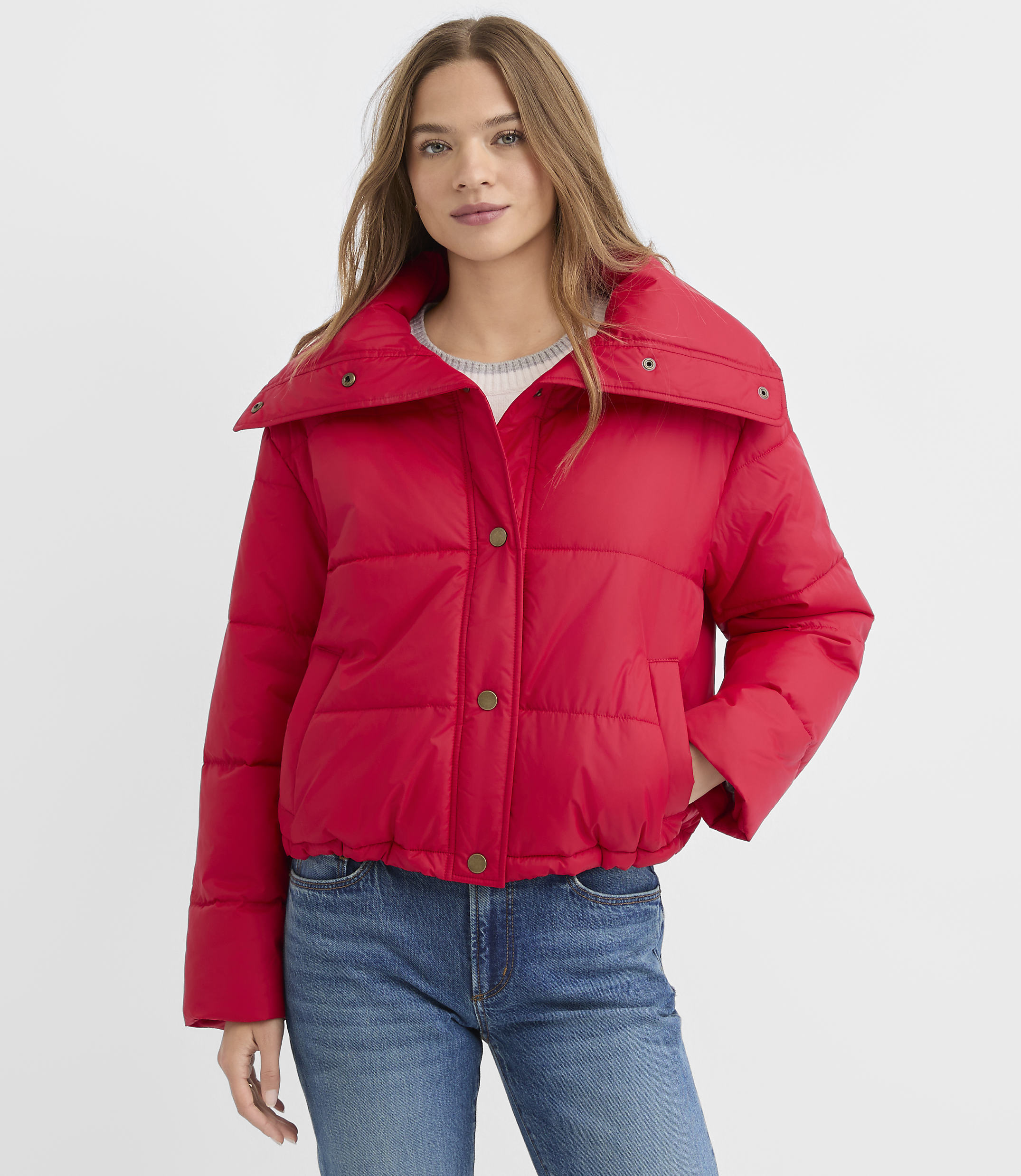 A woman wearing a bright red puffer jacket and blue jeans stands against a plain white background.