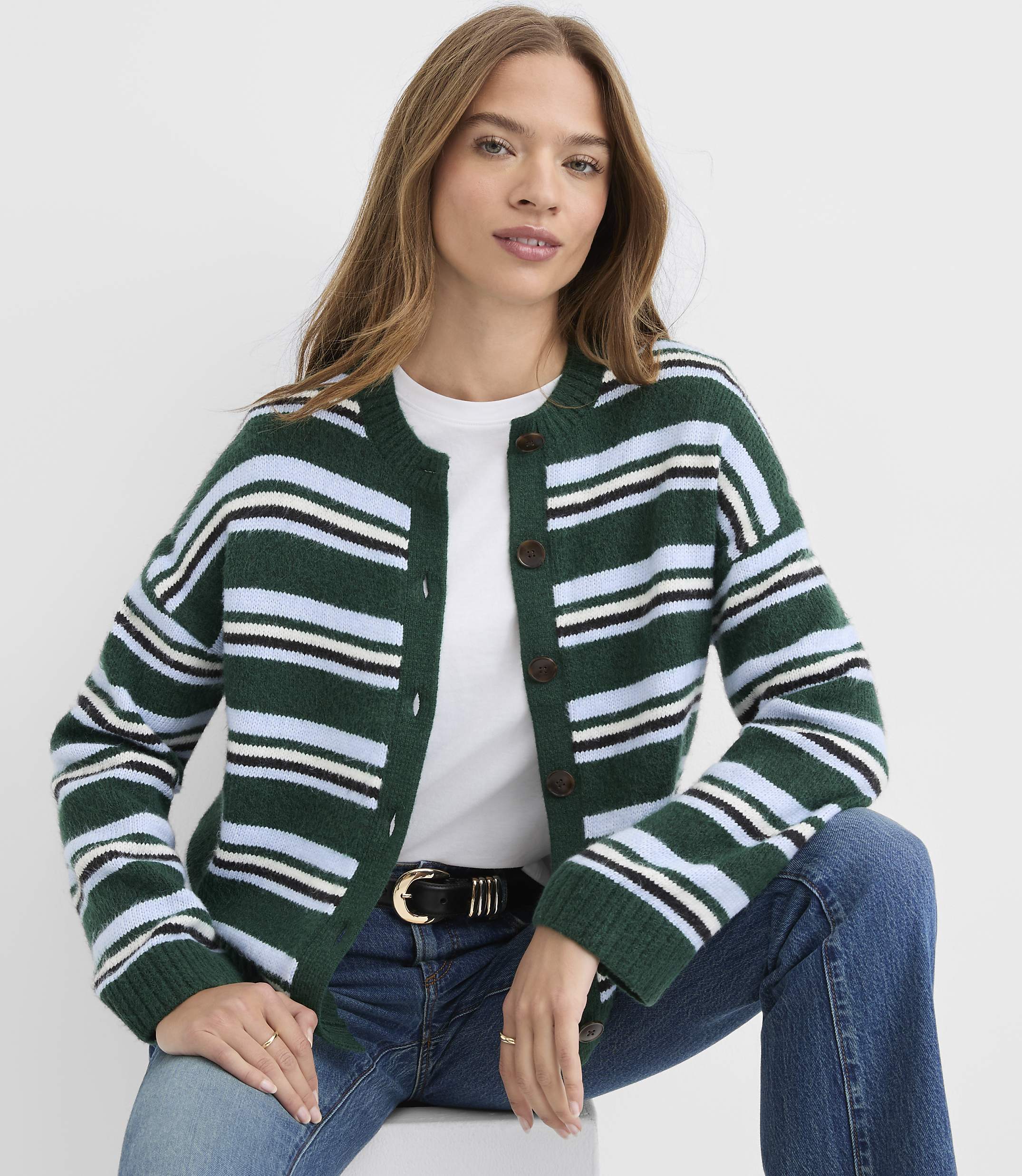 A woman wearing a striped green cardigan and blue jeans sits against a plain white background.