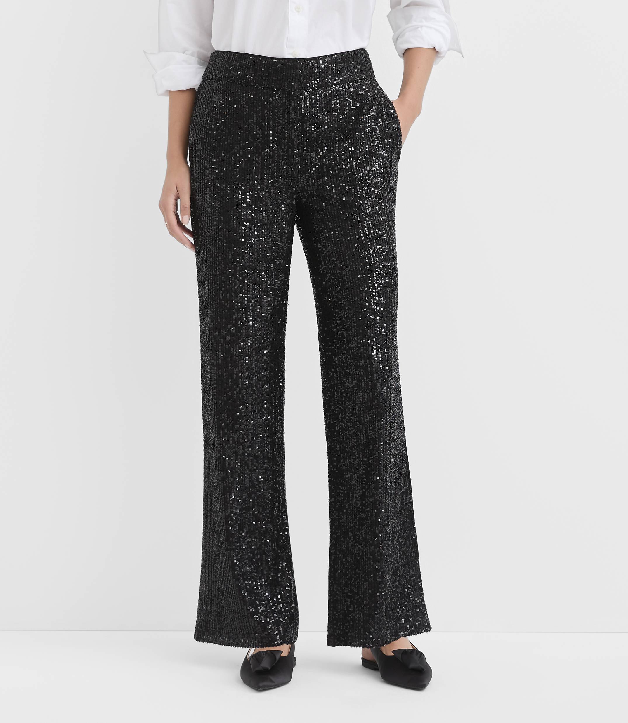 A person wears a white button-down shirt and shimmering black sequined trousers against a plain white background.
