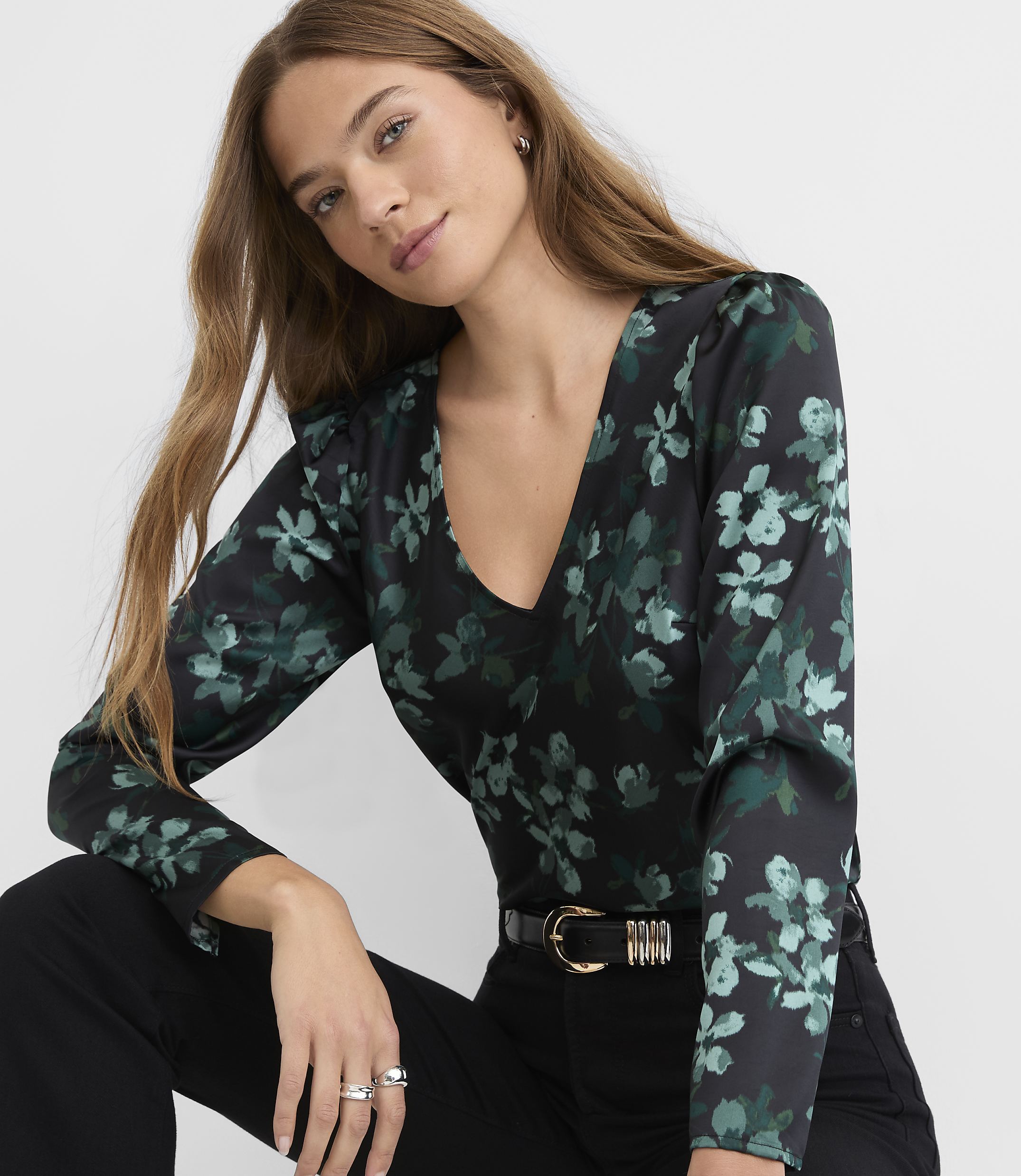 A woman wearing a black and green floral print blouse and black pants poses against a plain white background.