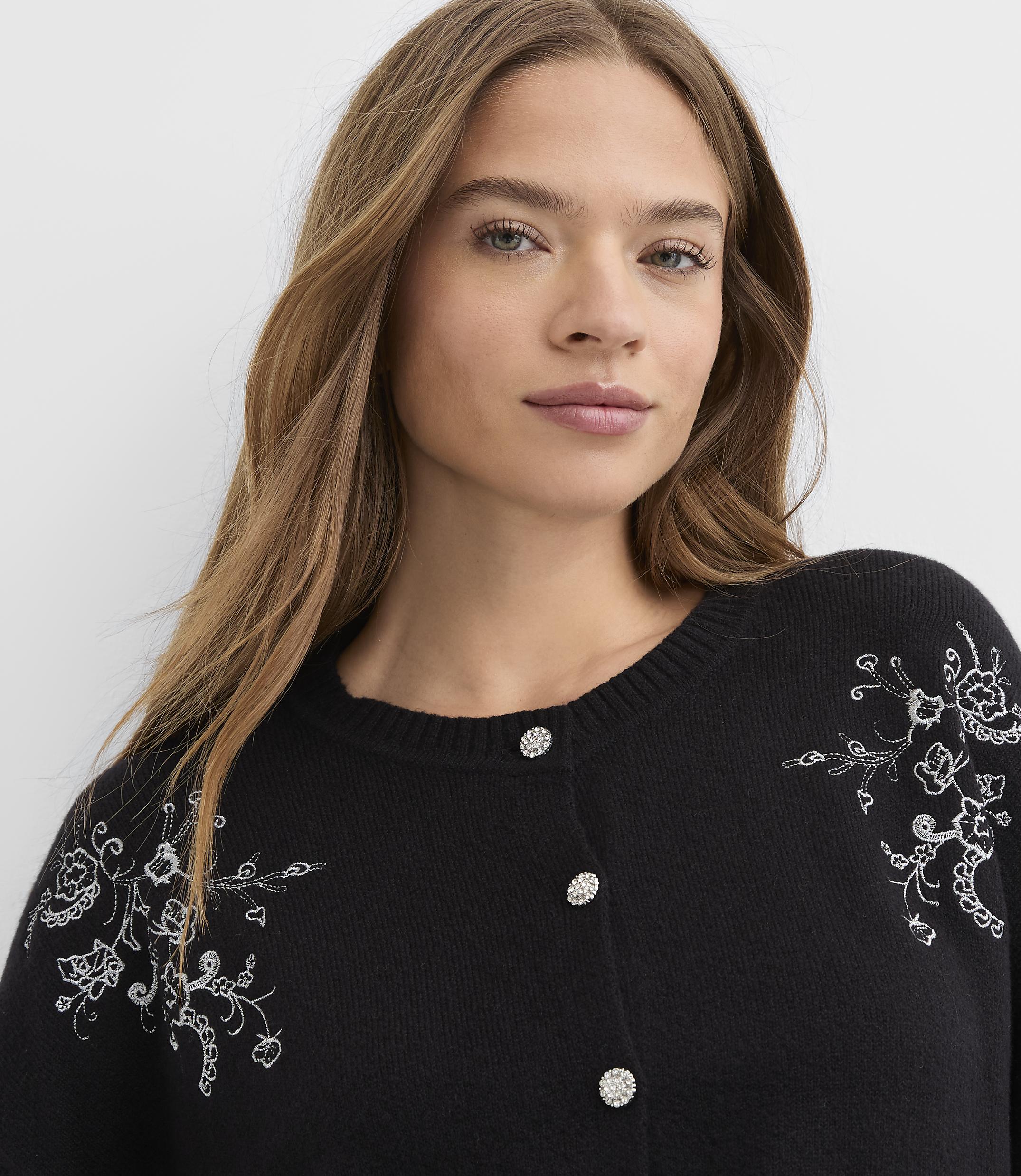 A woman with long brown hair wearing a black embroidered cardigan stands against a plain white background.