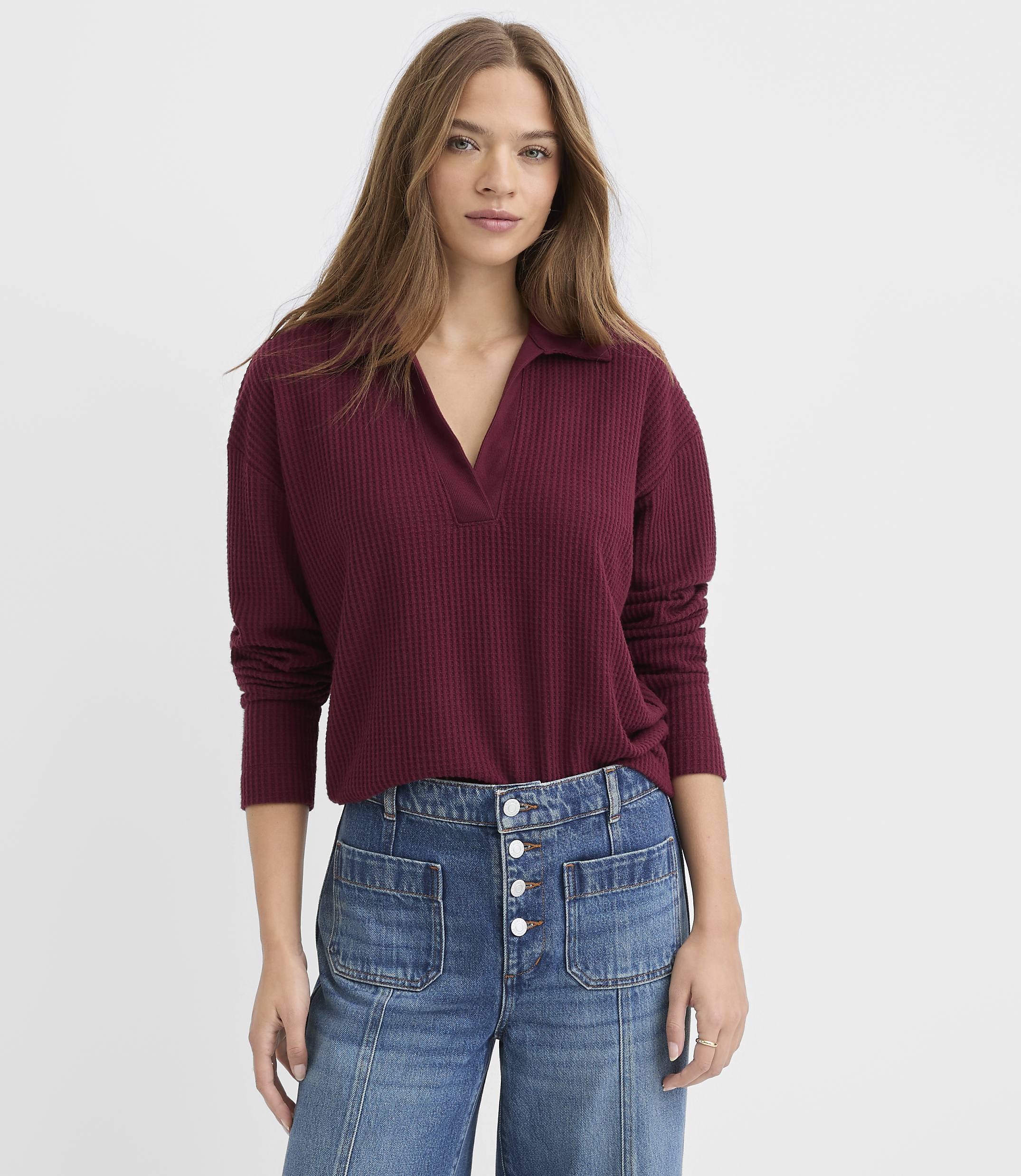 A woman wearing a ribbed burgundy v-neck top and high-waisted blue jeans stands against a plain white background.