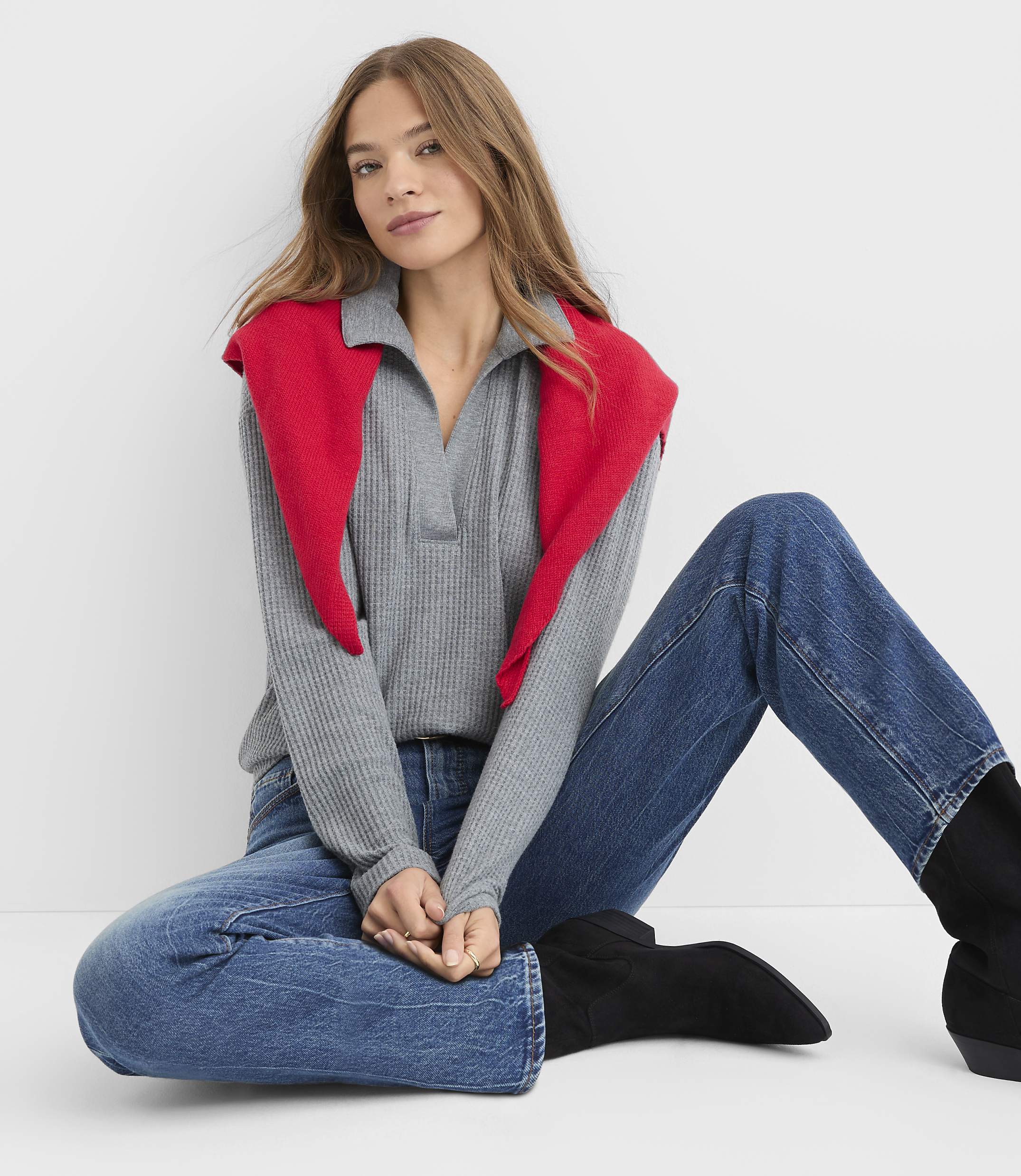 A woman wearing a grey ribbed top with a red shawl and blue jeans sits on the floor against a plain white background.
