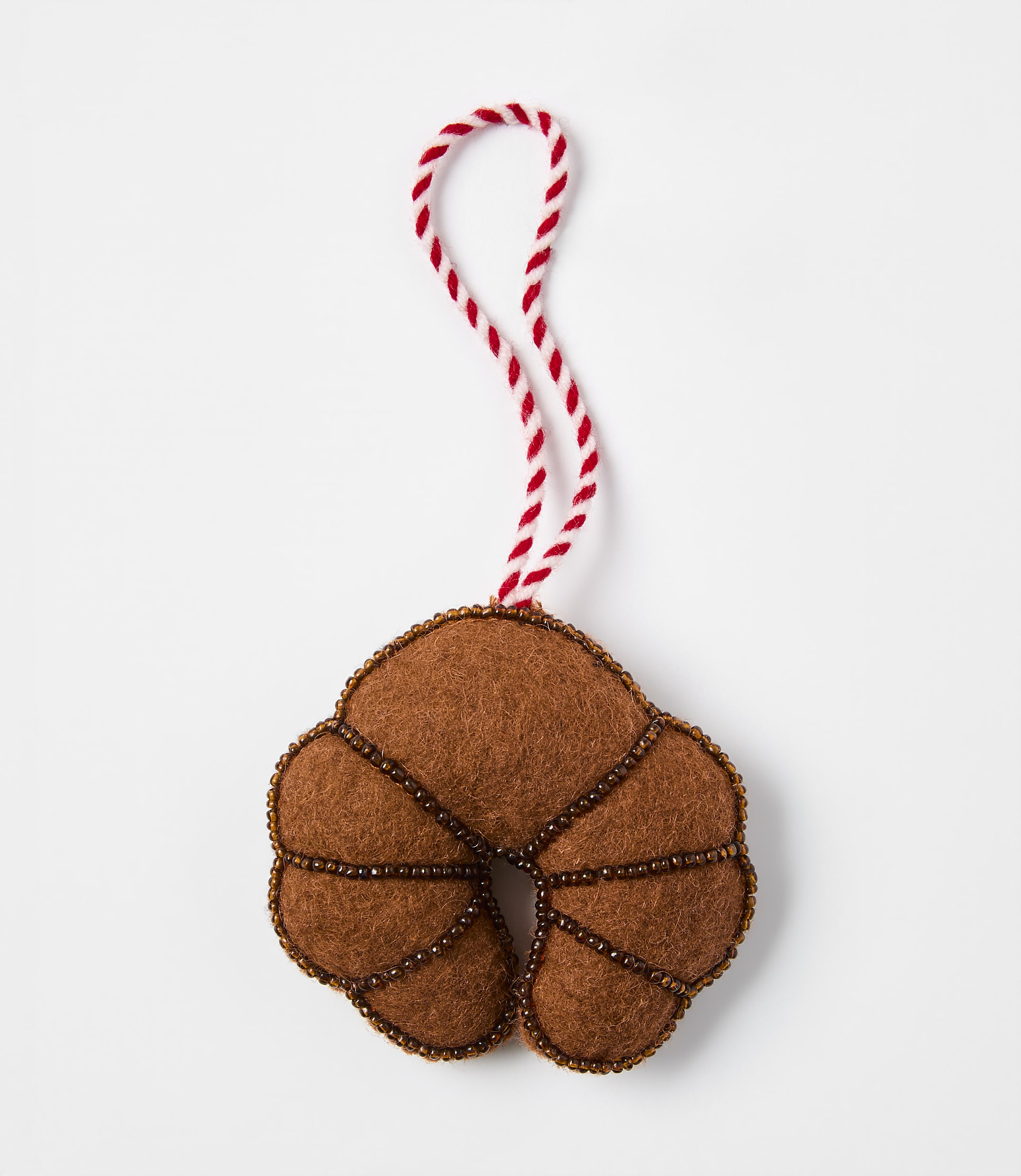 A brown felt croissant ornament with beaded detailing and a red-and-white string loop rests on a plain white background.