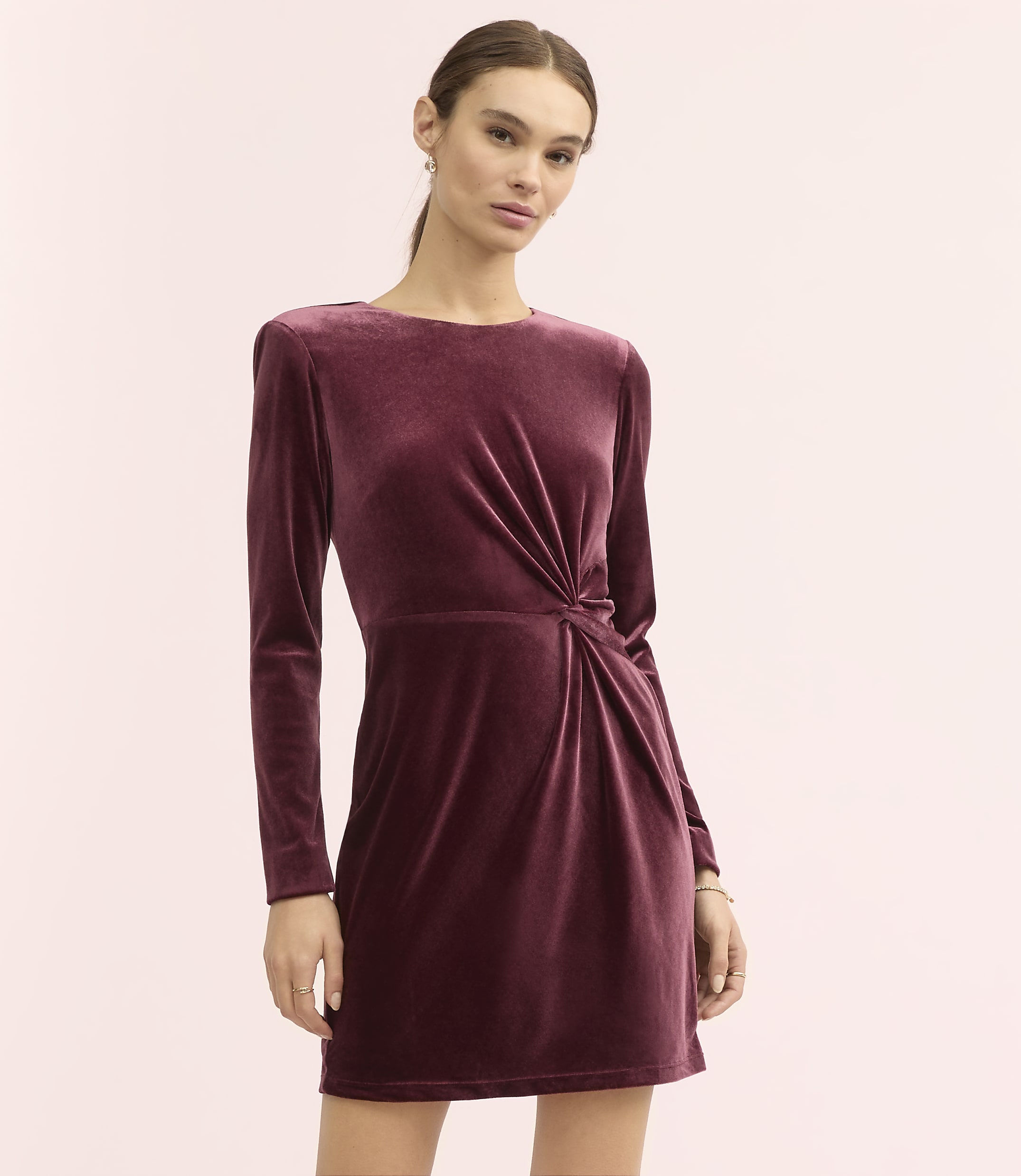 A woman wearing a long-sleeved, burgundy velvet mini dress stands against a plain, light pink background.