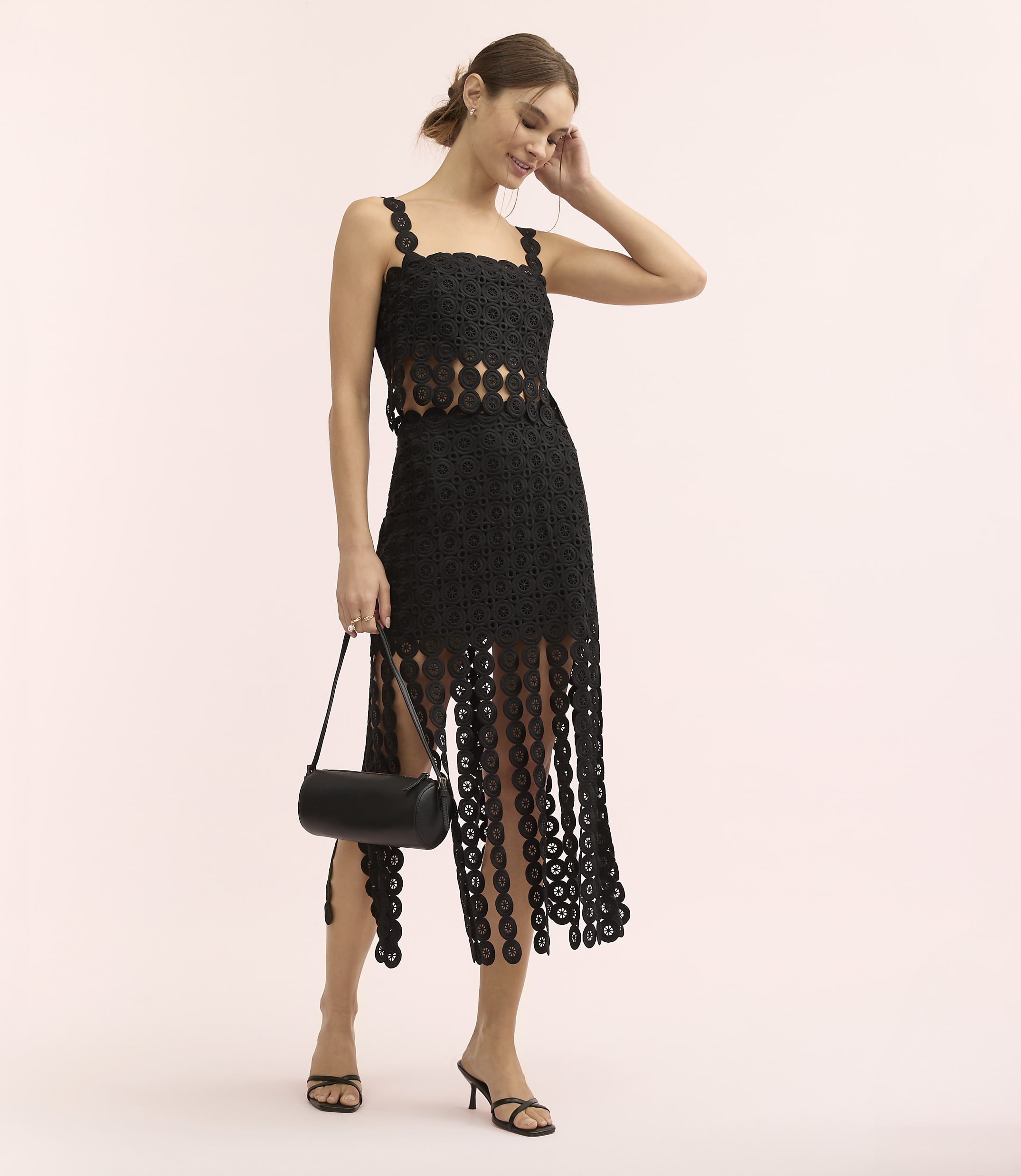 A woman wearing a black lace dress and holding a black handbag stands against a plain, light pink background.