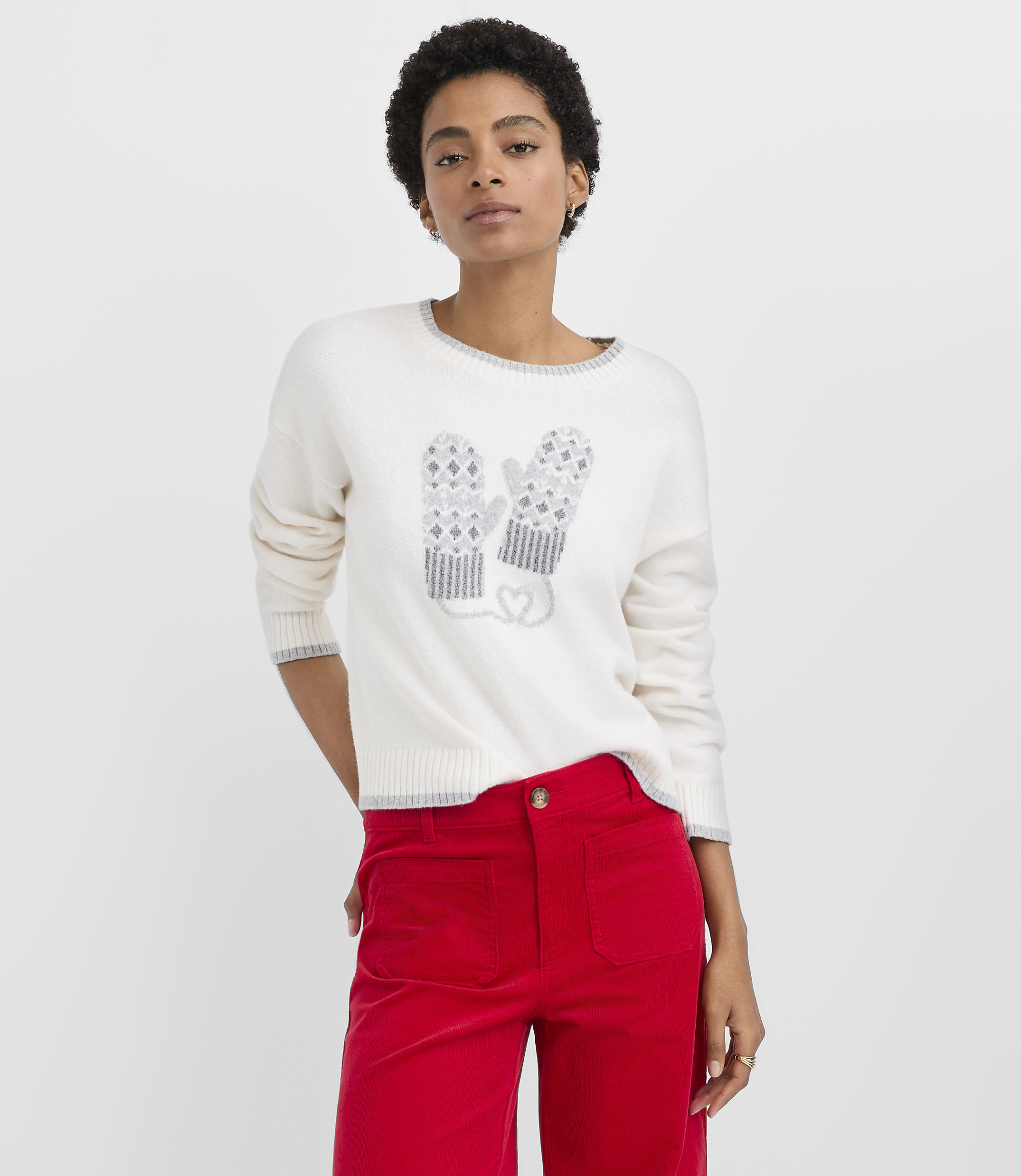 A woman wearing a white sweater with a mitten design and red pants stands against a plain white background.