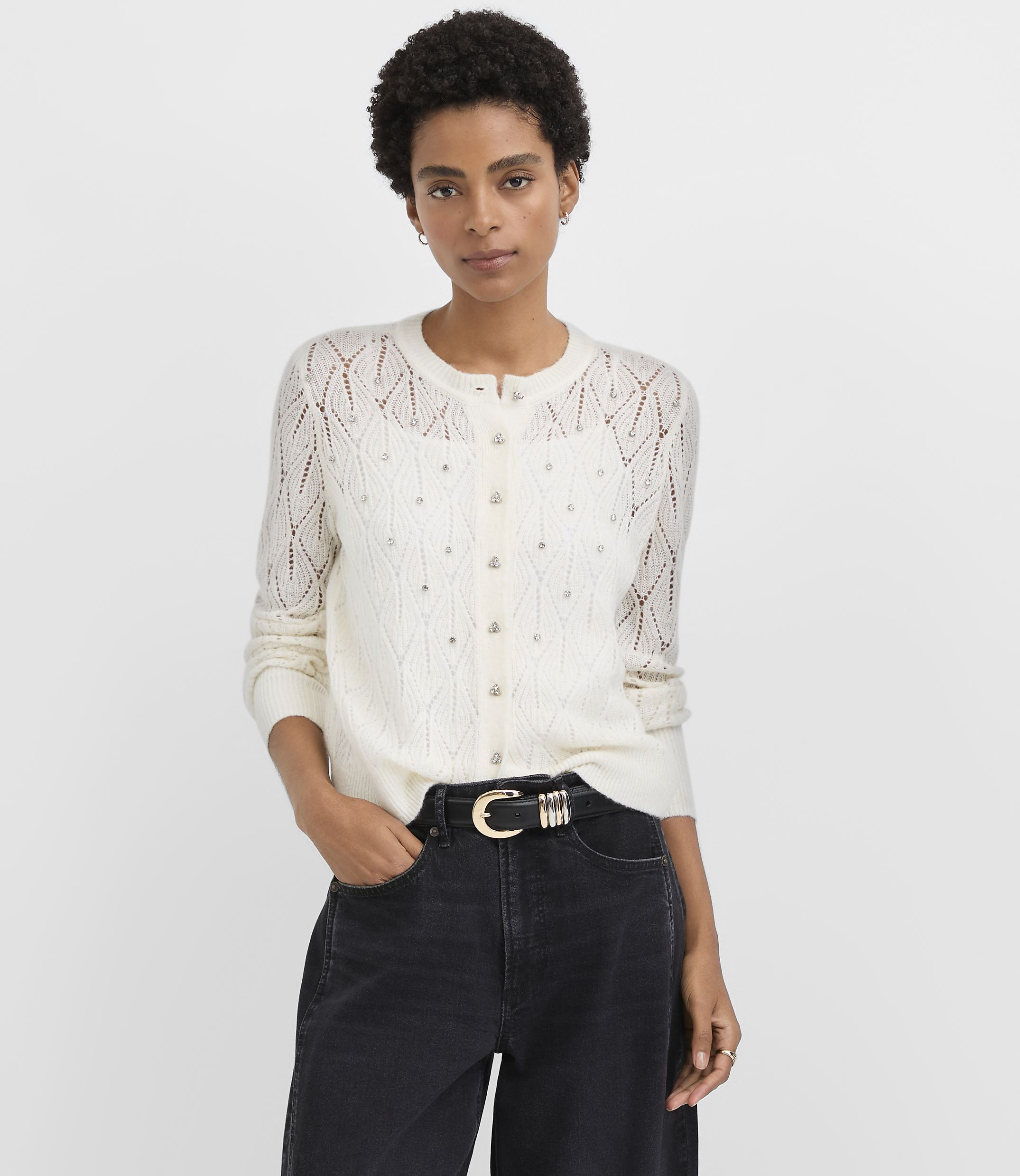 A woman wearing a white pointelle knit cardigan and black jeans stands against a plain light gray background.