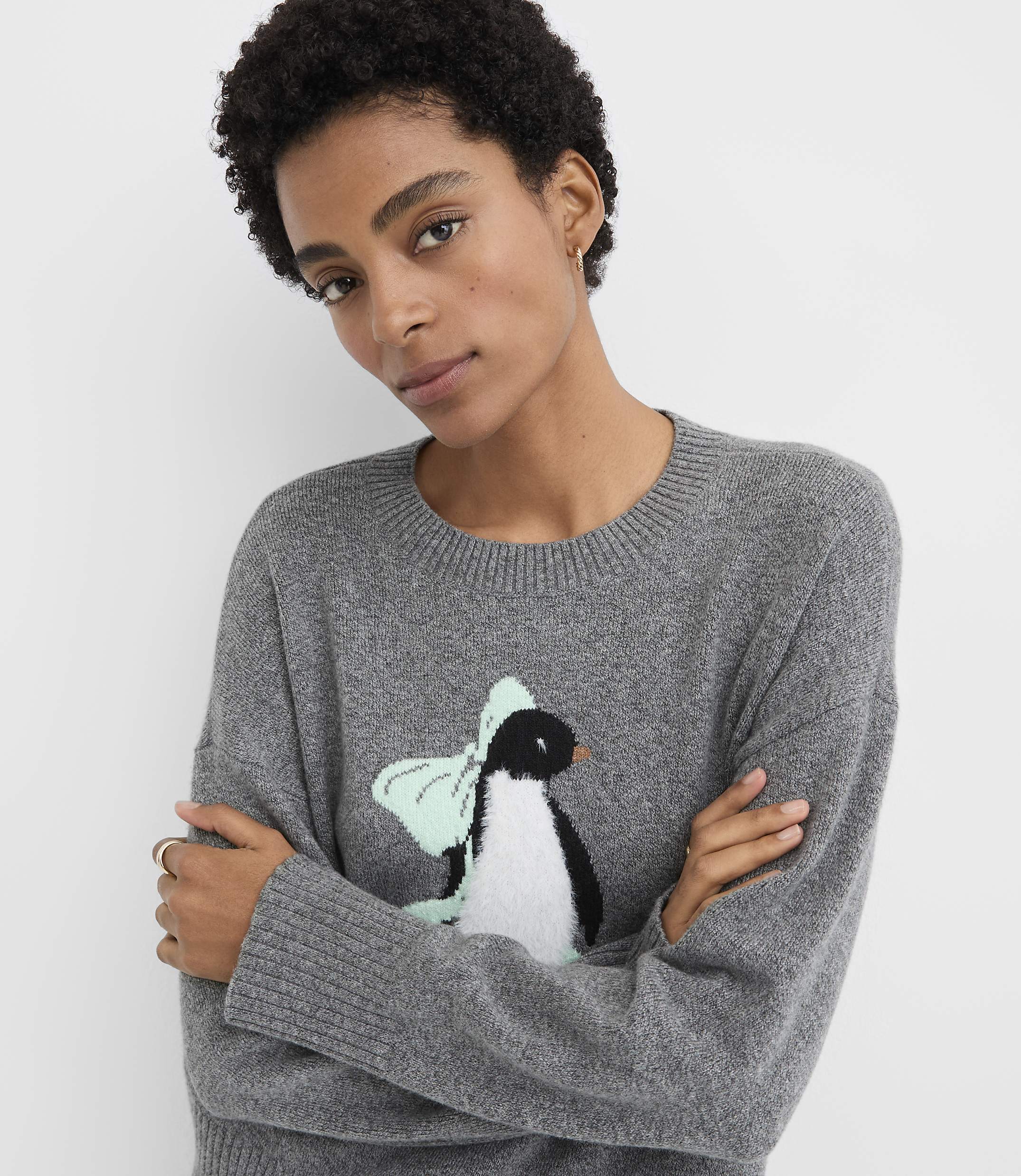A woman with short curly hair wearing a grey sweater with a penguin graphic stands against a plain white background.
