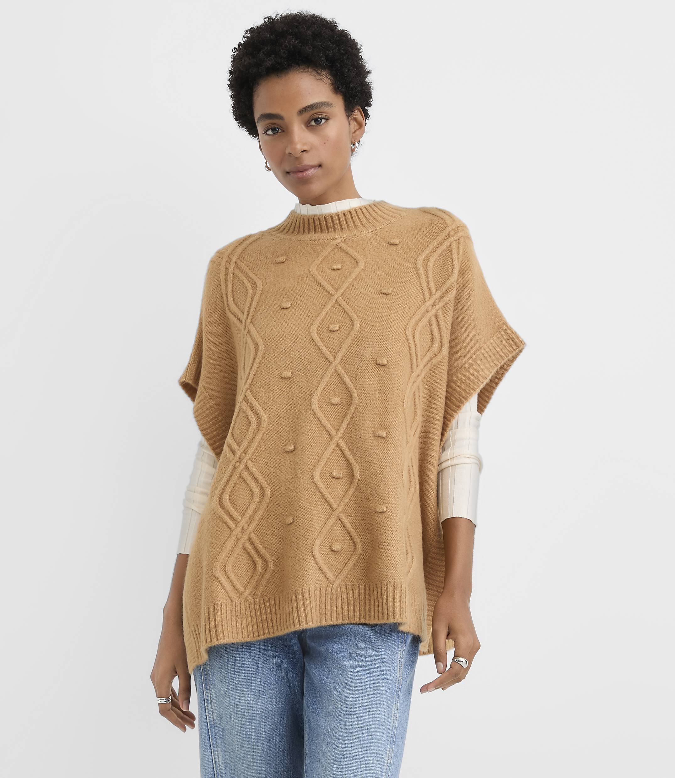 A woman wearing a tan cable-knit poncho and blue jeans stands against a plain white background.