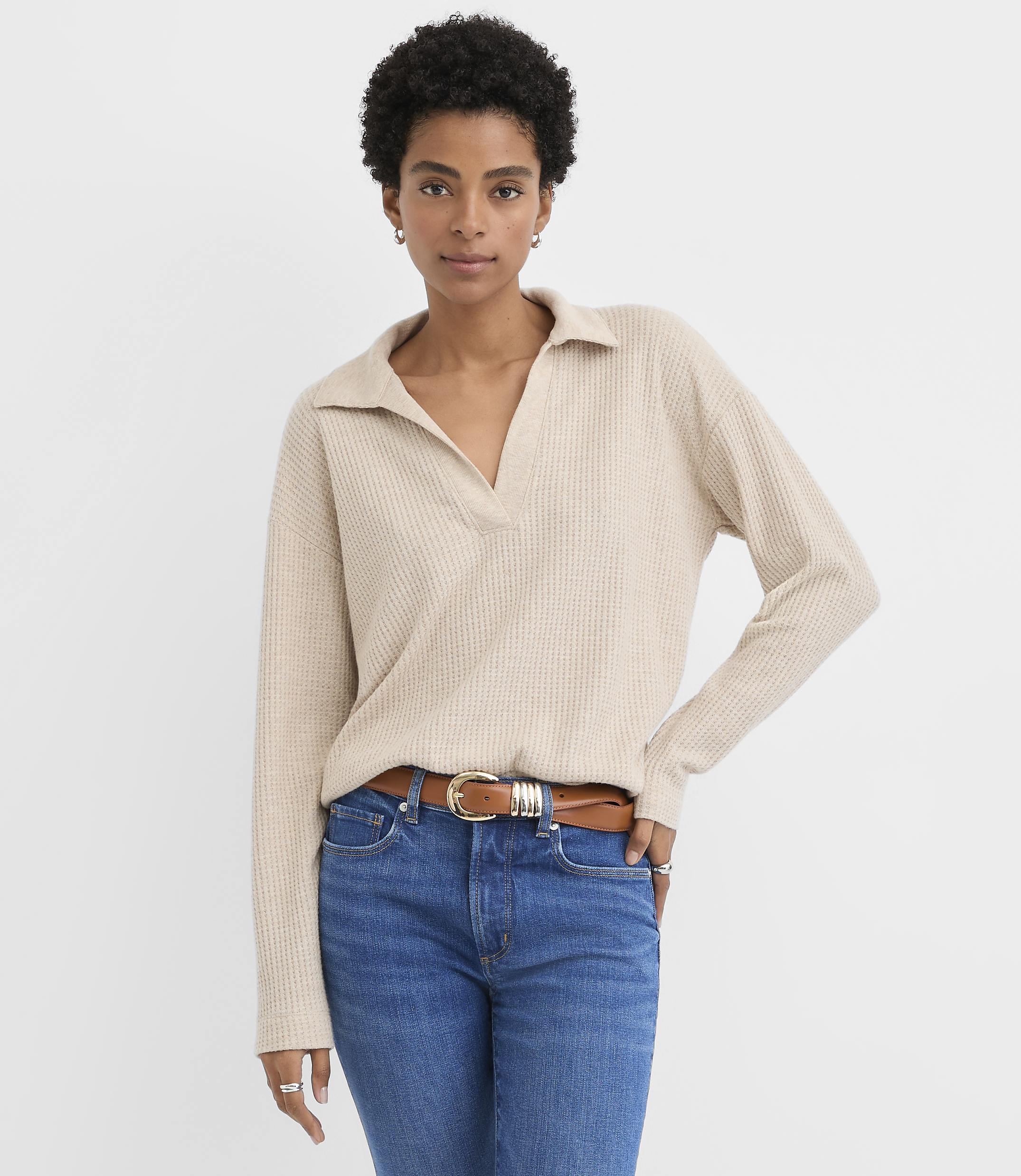 A woman wearing a beige collared sweater and blue jeans stands against a plain white background.