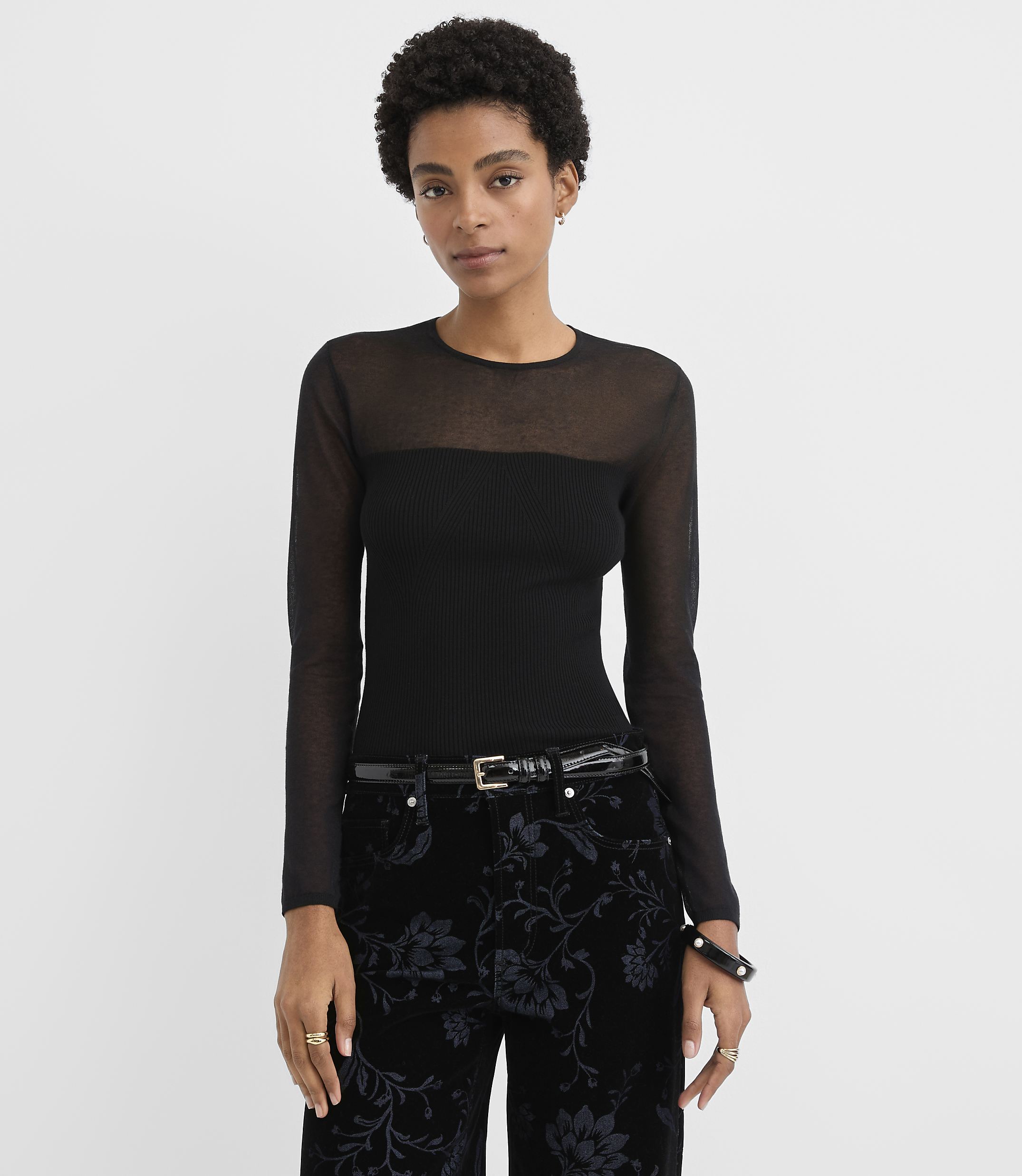A woman wearing a sheer-sleeved black top and floral patterned black pants stands against a plain white background.