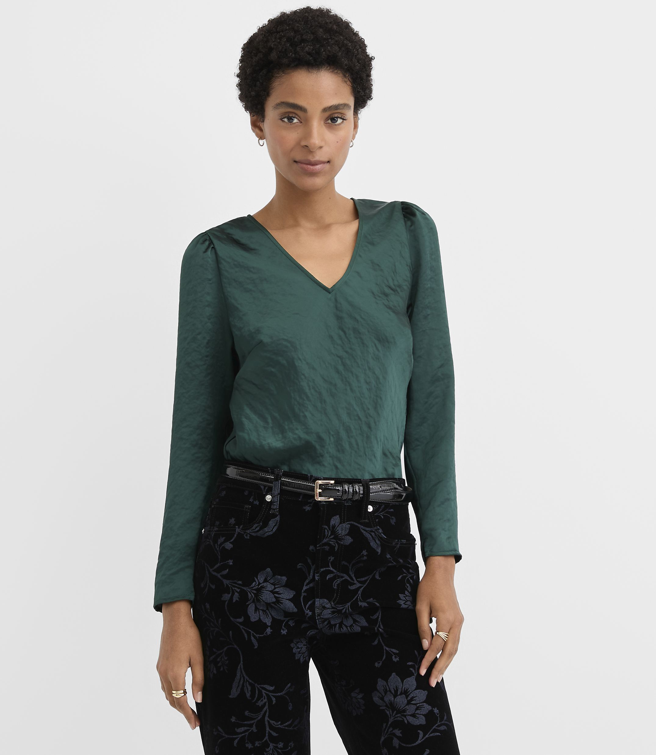 A woman wearing a dark green V-neck blouse and floral black trousers stands against a plain white background.