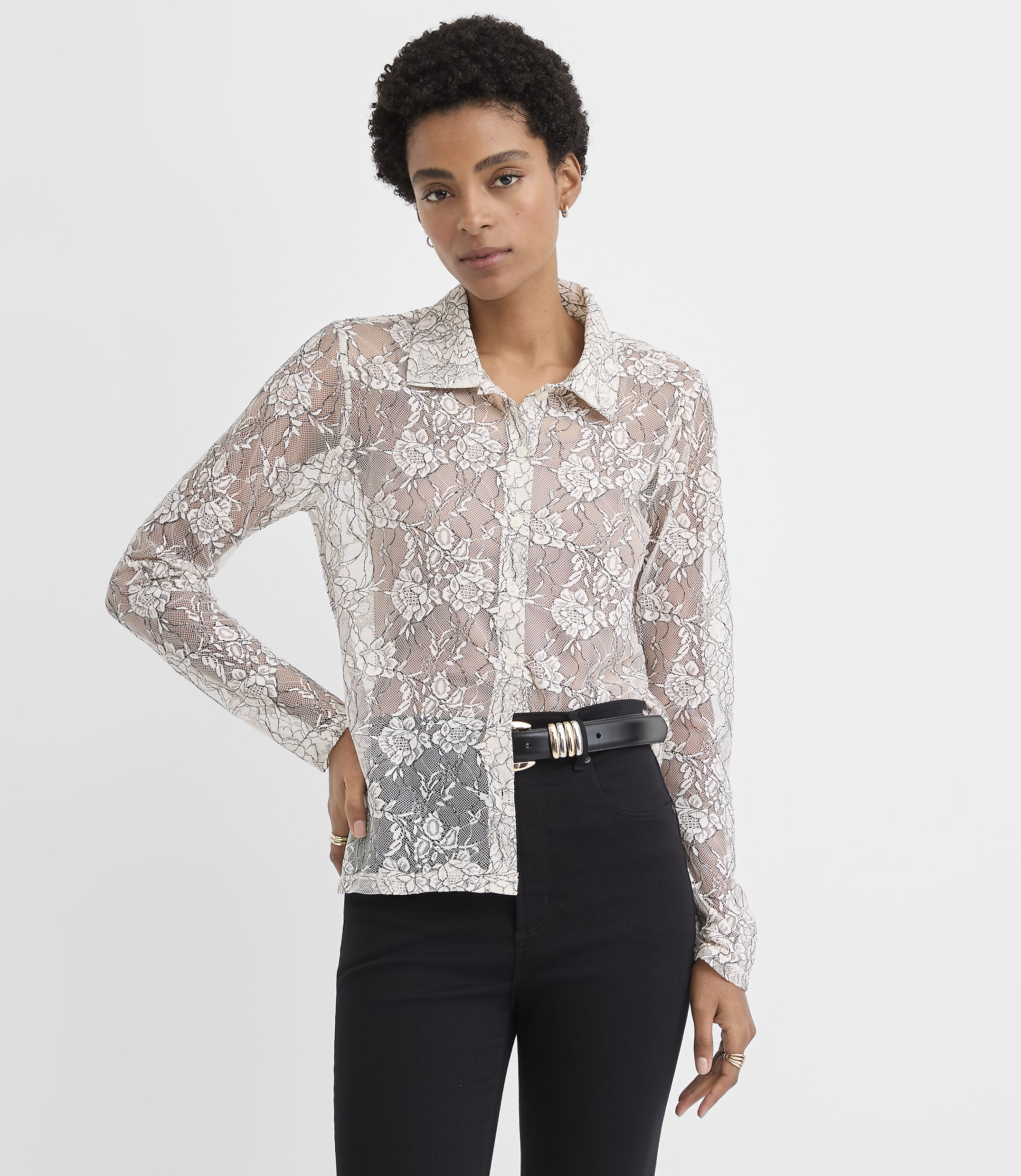 A woman wearing a sheer white lace button-down shirt and black trousers stands against a plain white background.