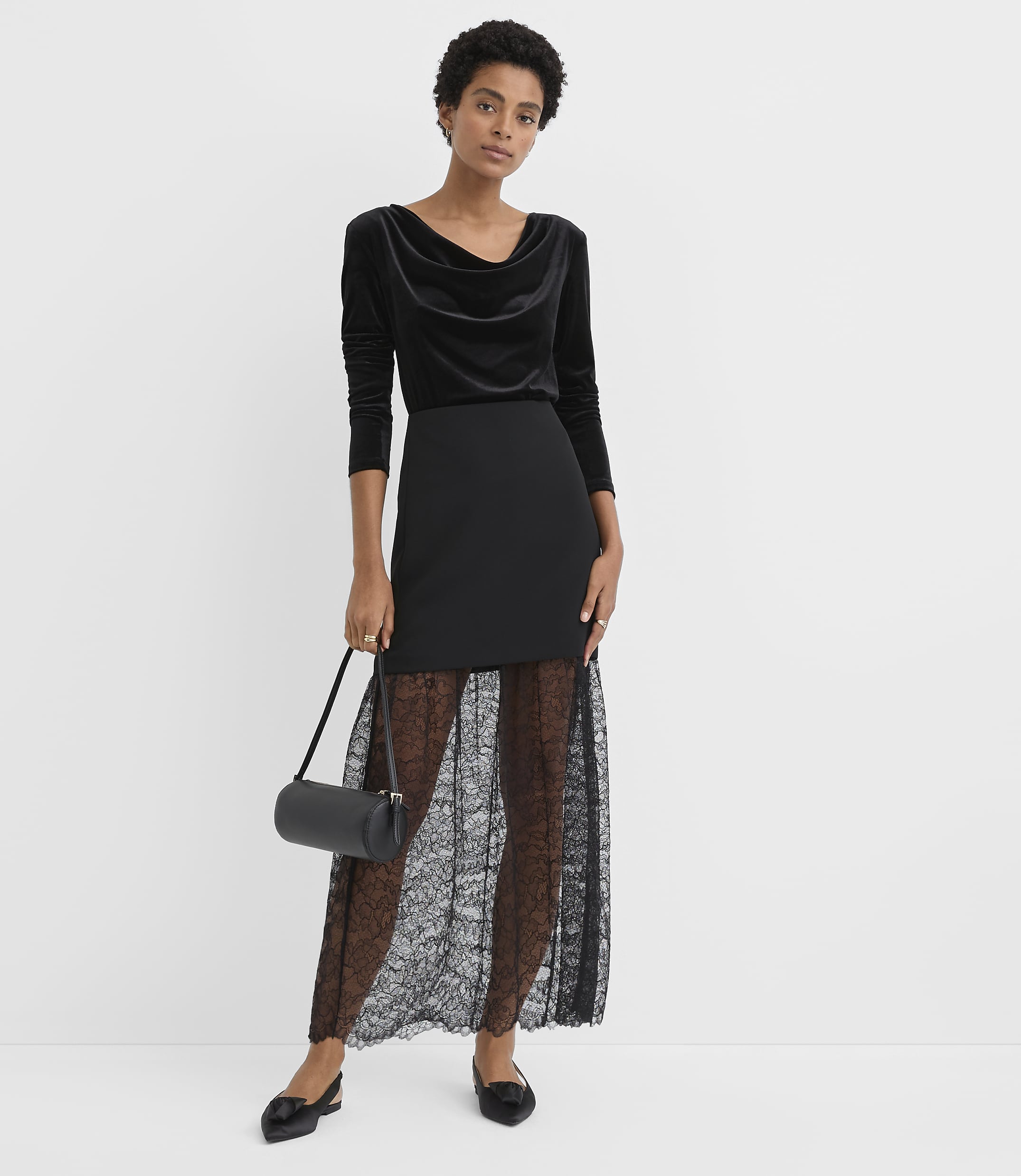 A woman wearing a black velvet top and a black lace skirt stands against a plain white background.