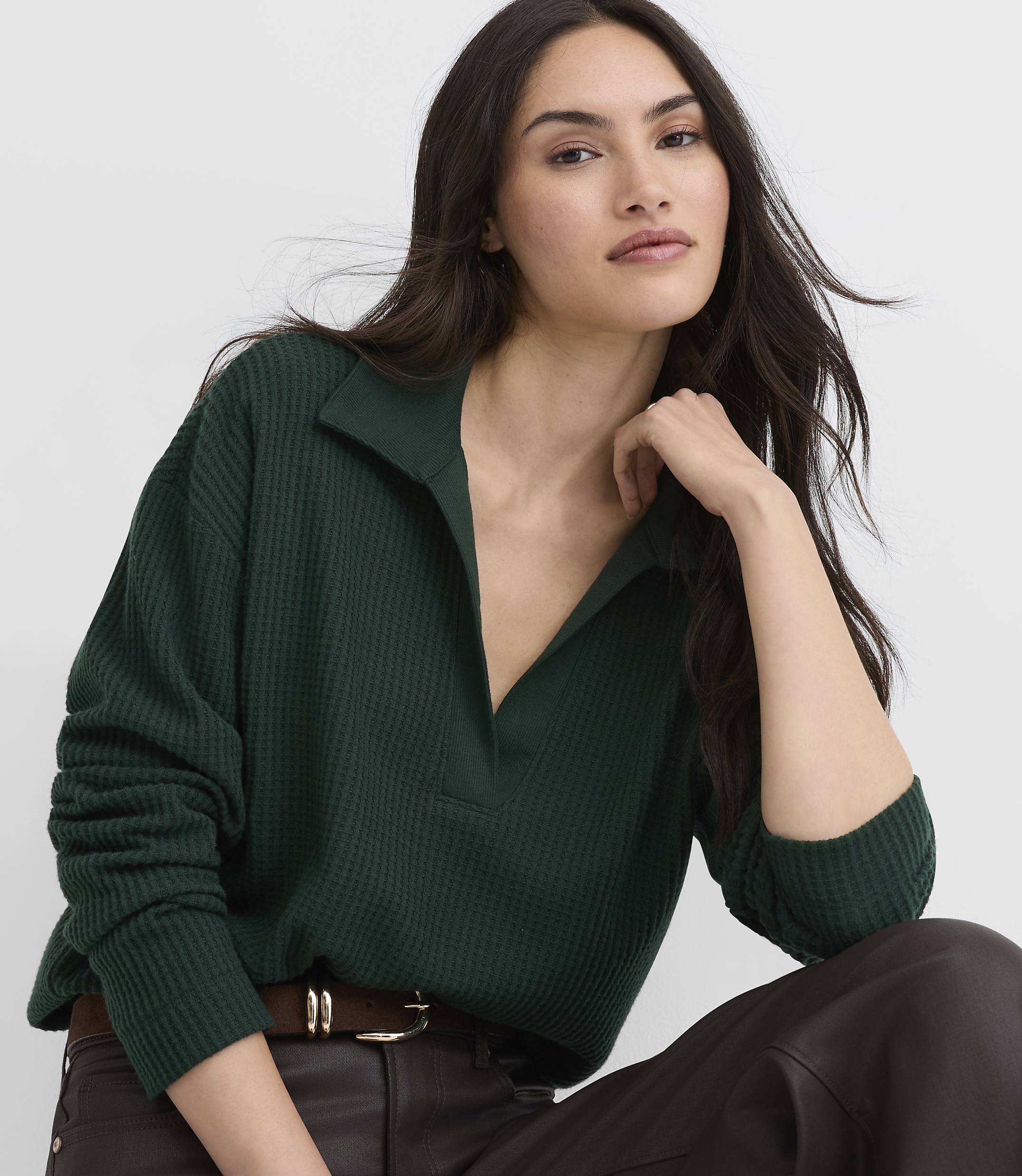 A woman wearing a dark green ribbed polo shirt and brown leather pants poses against a plain white background.