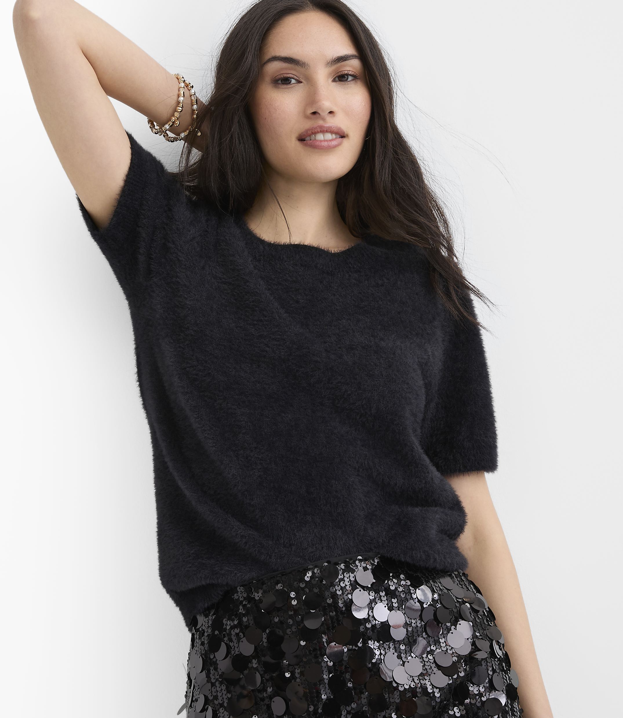 A woman wearing a fuzzy black top and a sequined skirt poses against a plain white background.