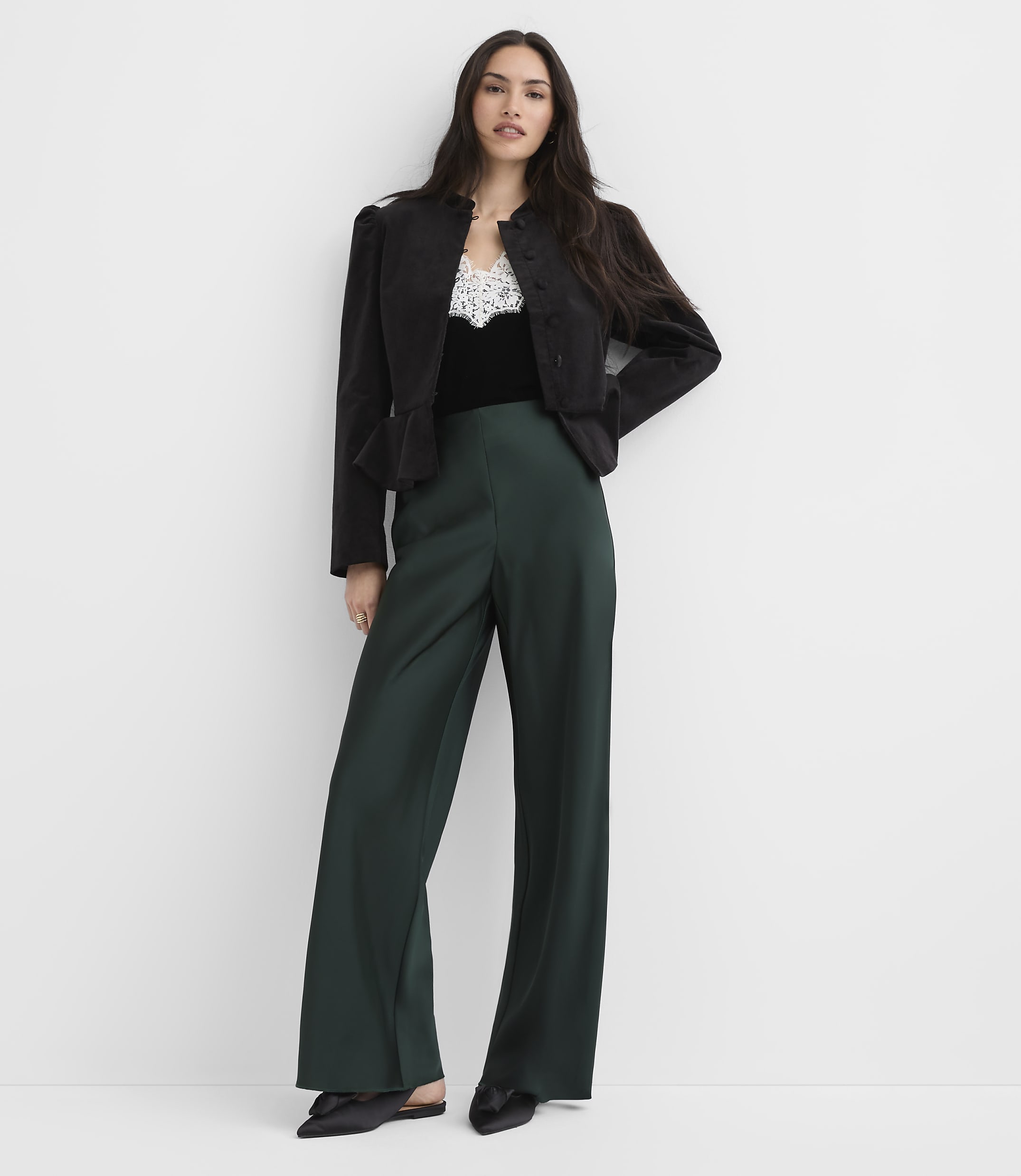 A woman wearing a black jacket, a lace-trimmed top, and wide-leg dark green trousers stands against a plain white background.