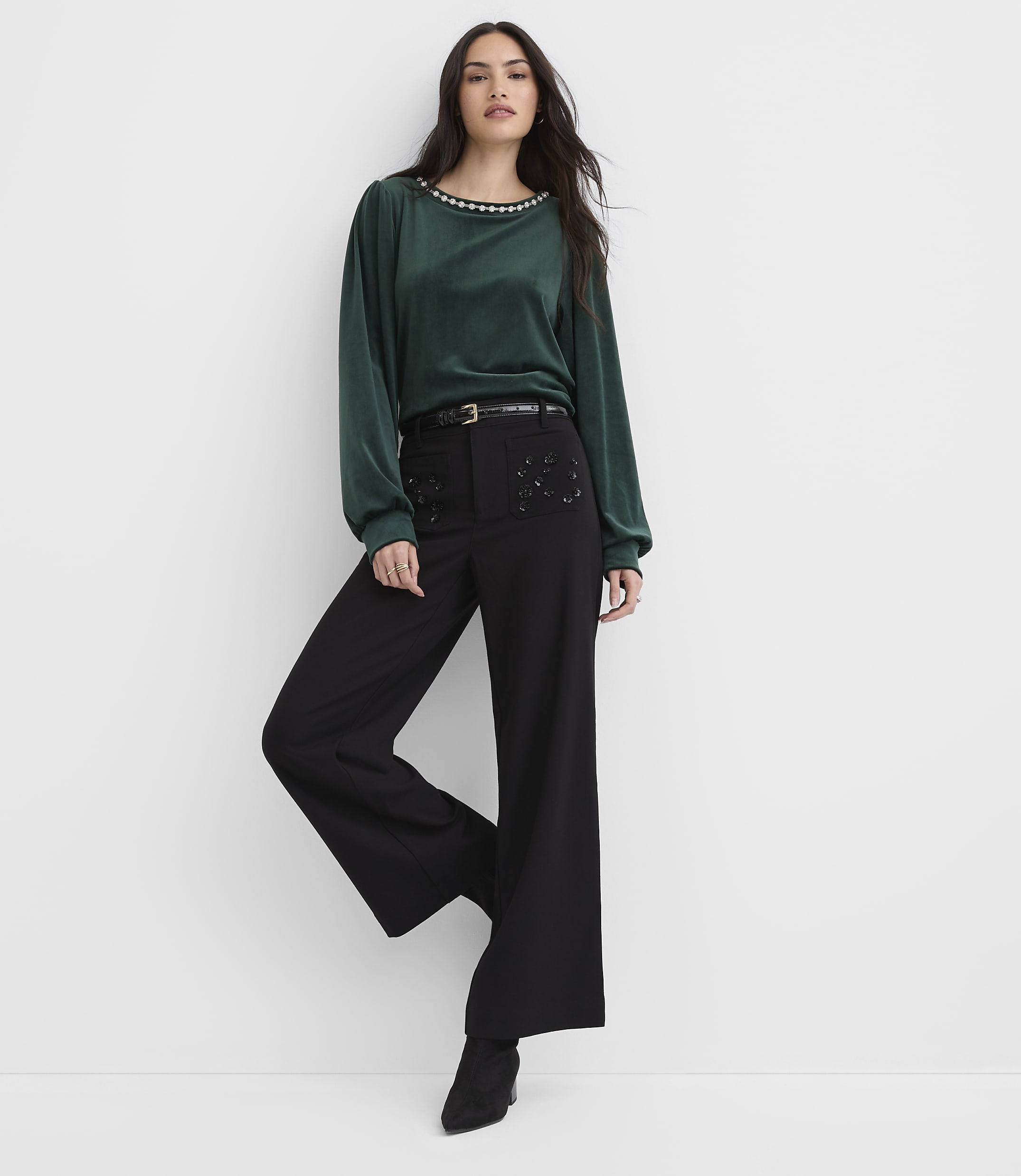 A woman wearing a green long-sleeved top and black wide-leg trousers stands against a plain white background.