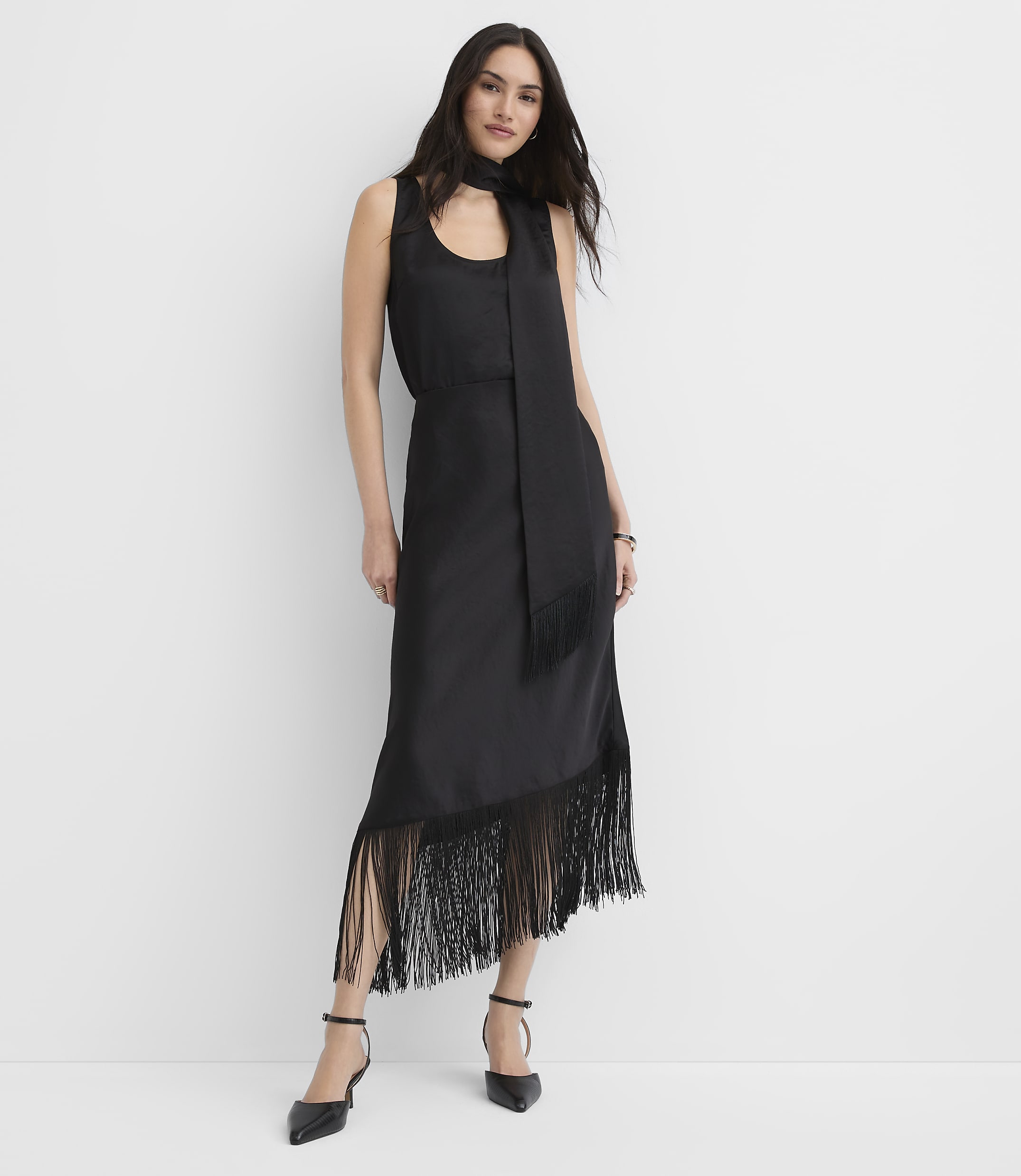 A woman wearing a black sleeveless midi dress with fringe detailing stands against a plain white background.