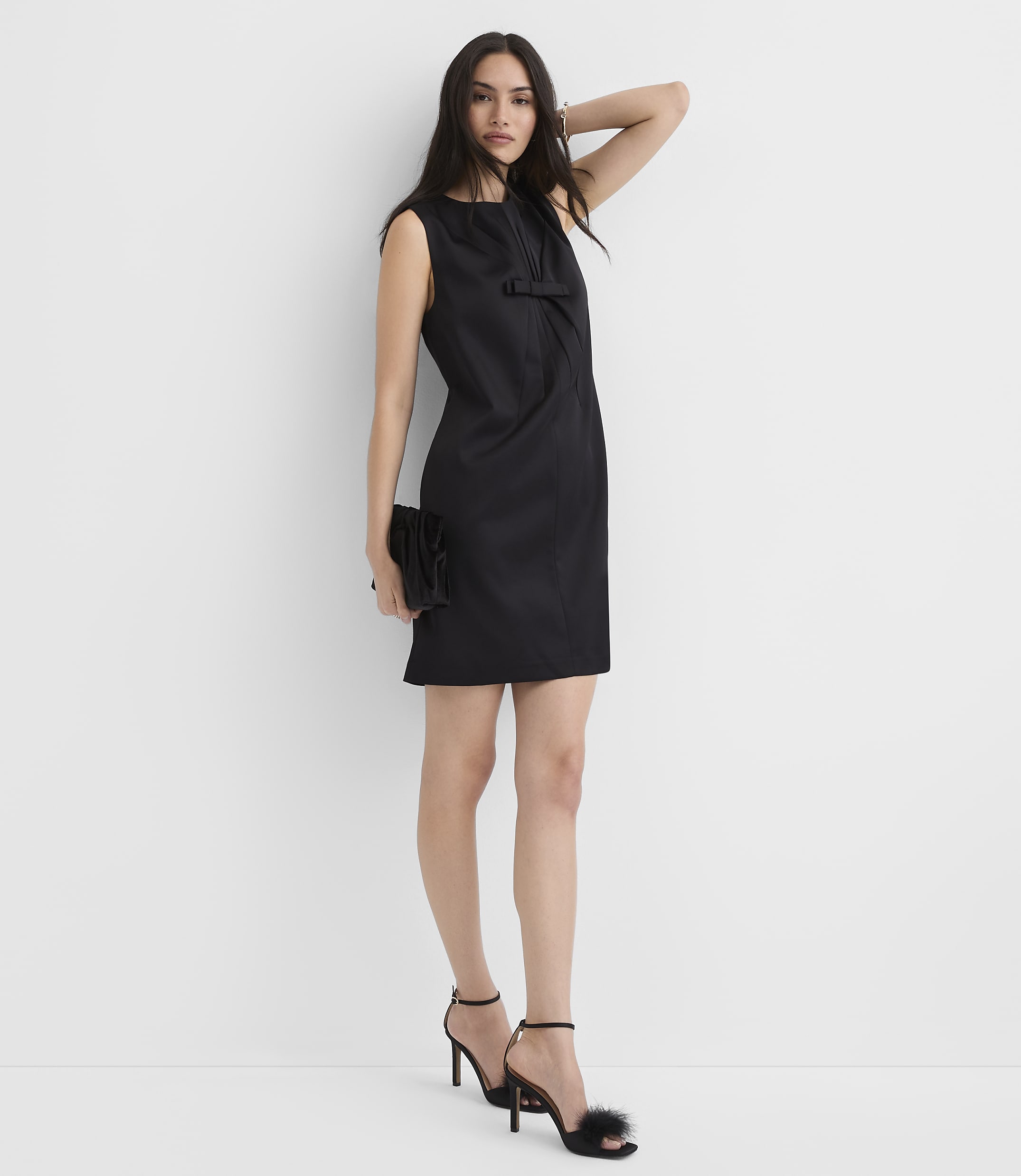 A woman wearing a short black dress and high-heeled sandals stands against a plain white background.