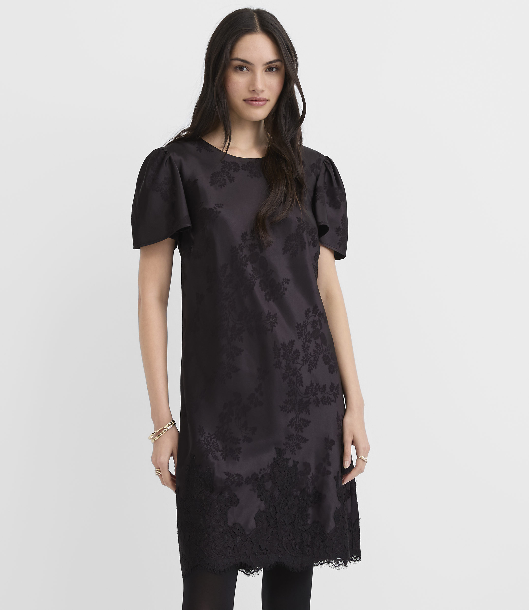 A woman wearing a short-sleeved black floral dress stands against a plain white background.