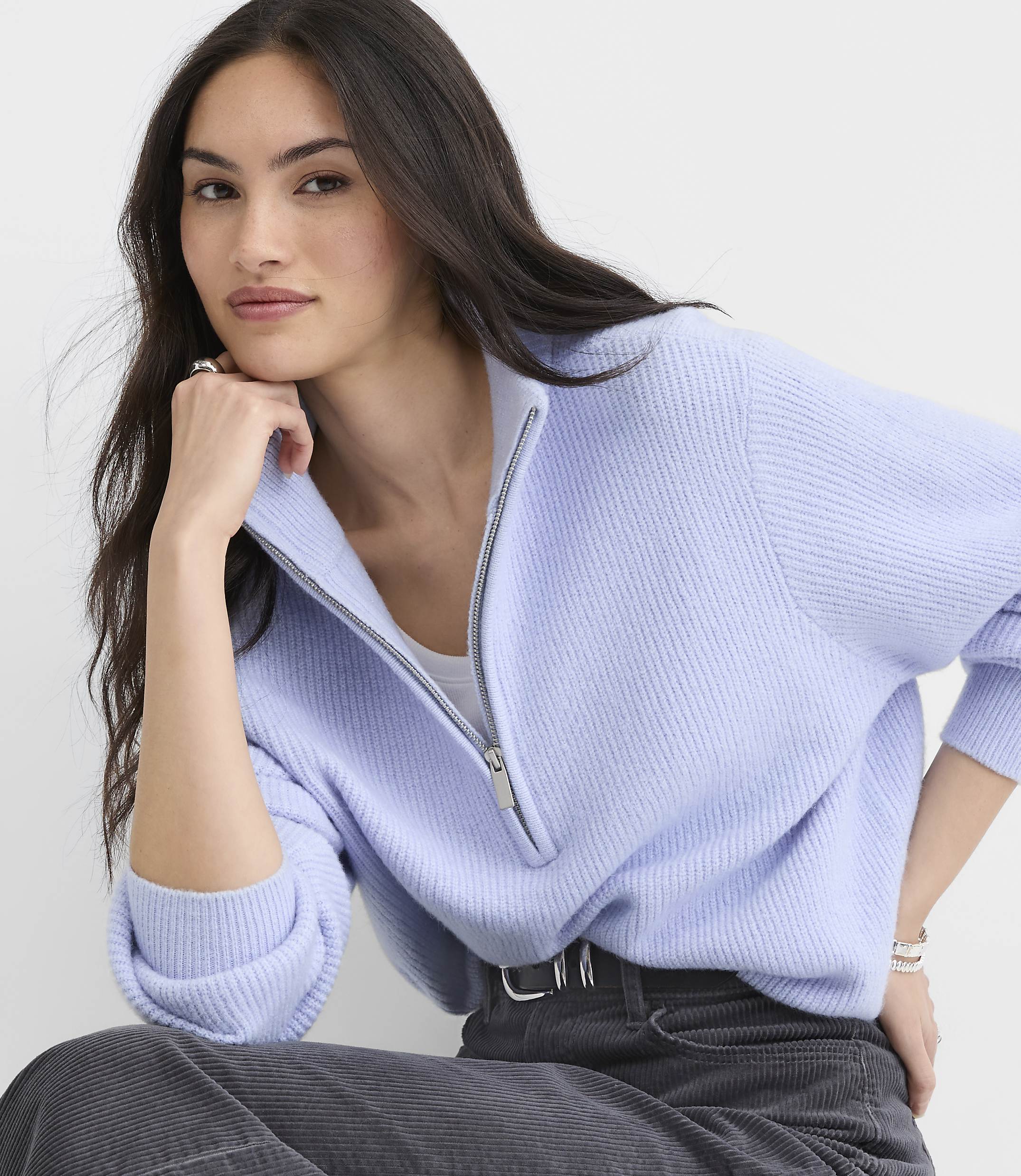 A woman wearing a light purple quarter-zip sweater and dark grey corduroy pants poses against a plain white background.