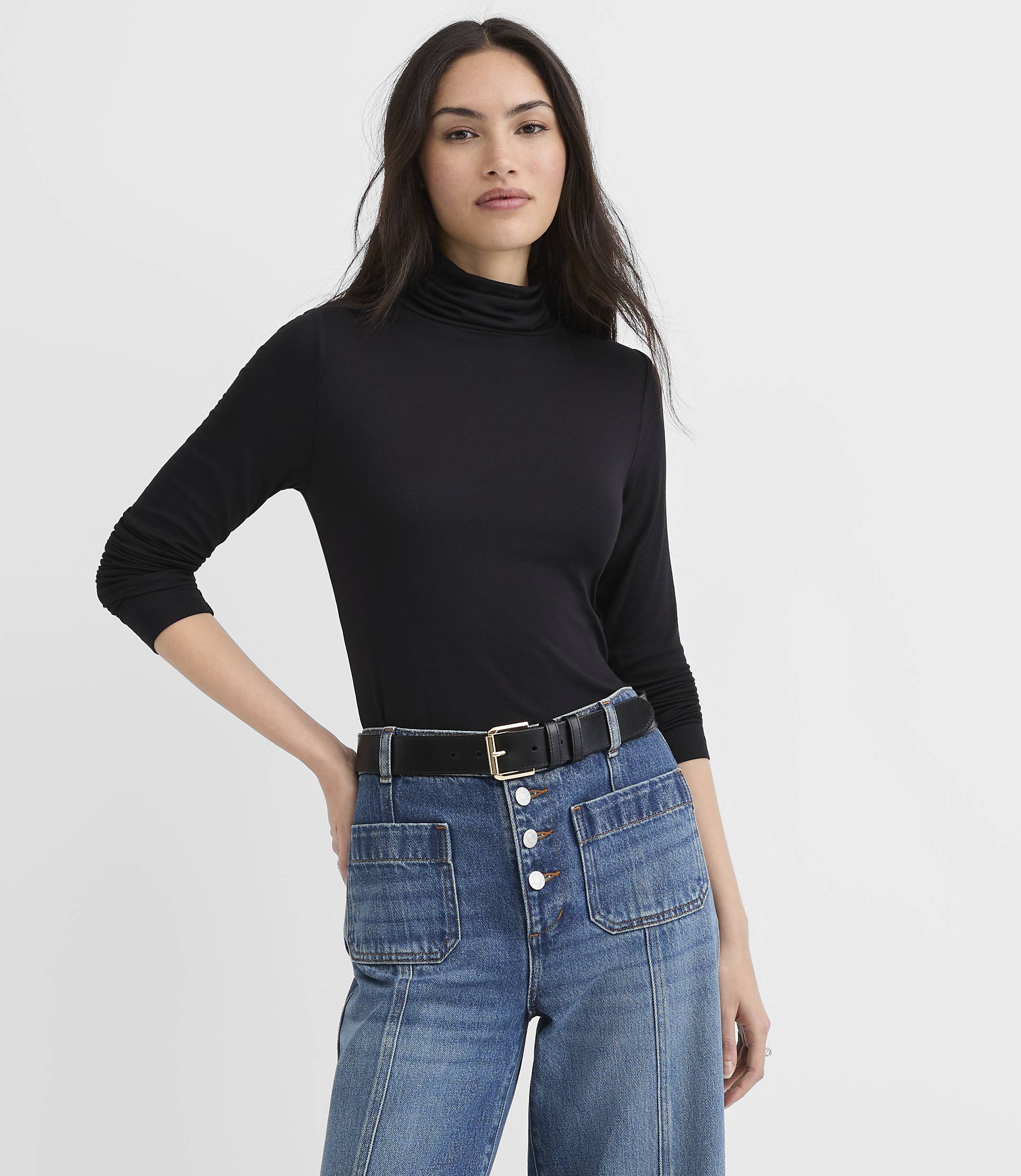 A woman wearing a black turtleneck and blue jeans stands against a plain white background.