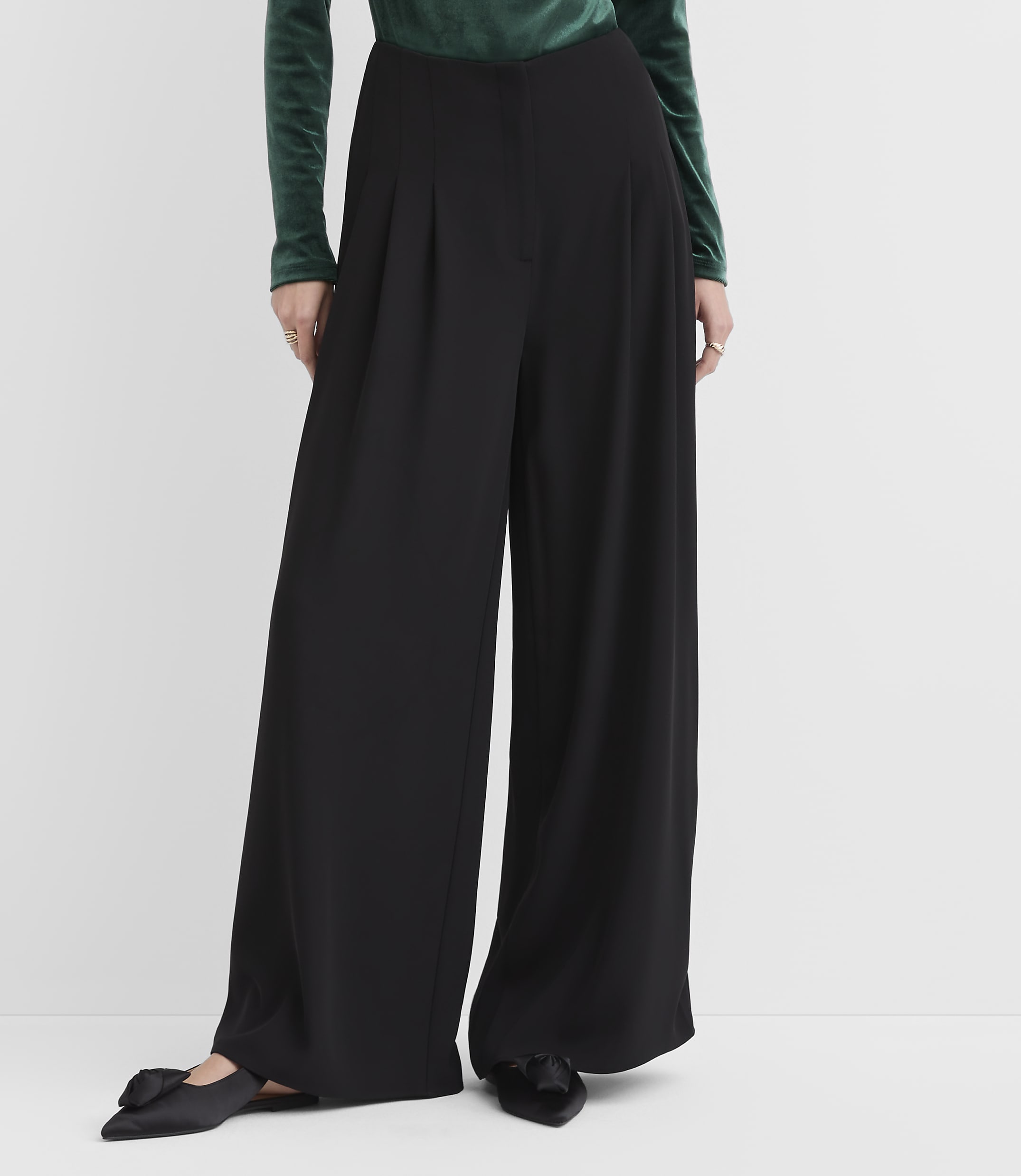 A person wearing a green velvet top and wide-leg black trousers stands against a plain white background.