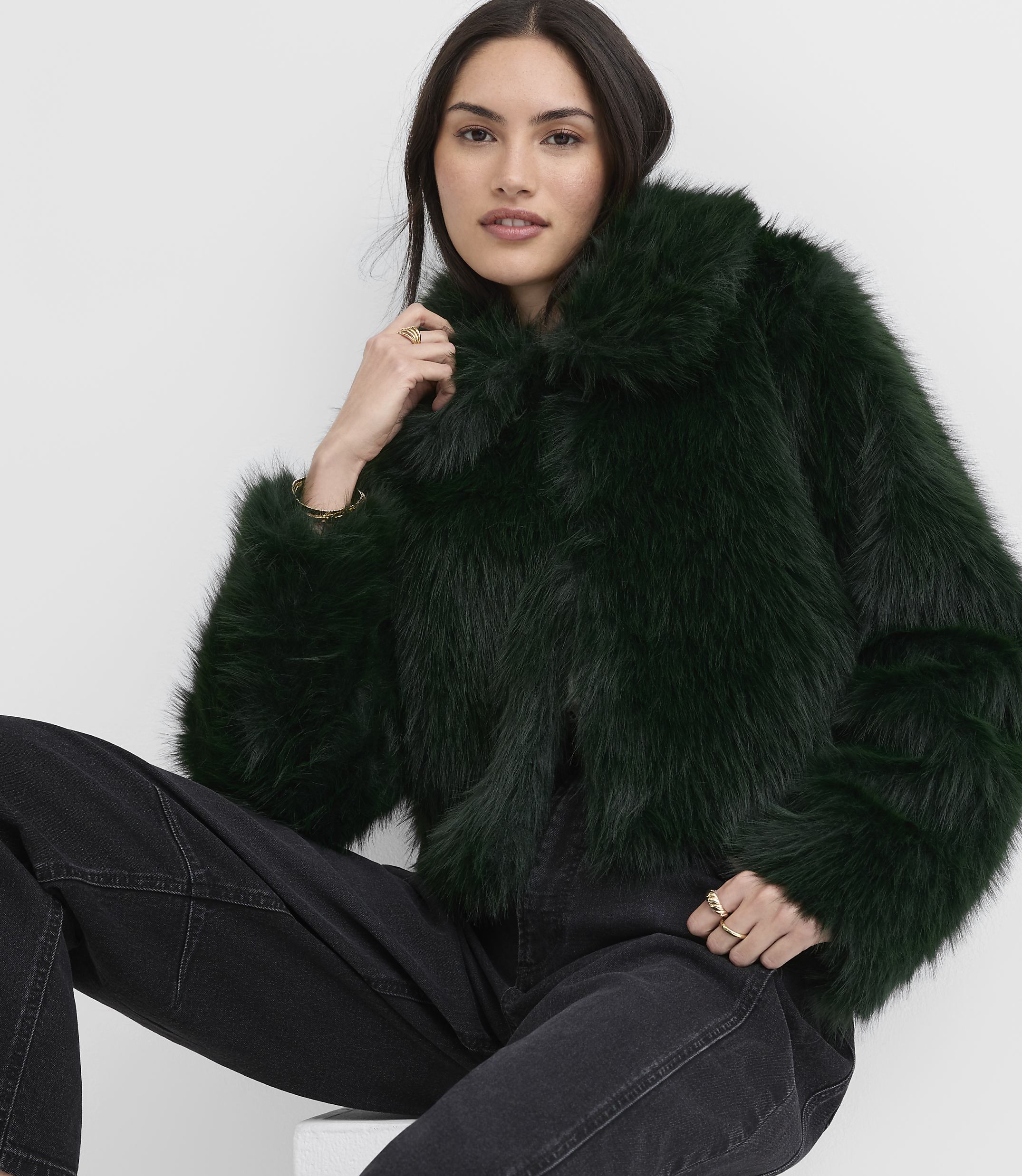 A woman wearing a dark green faux fur jacket and black jeans poses against a plain white background.