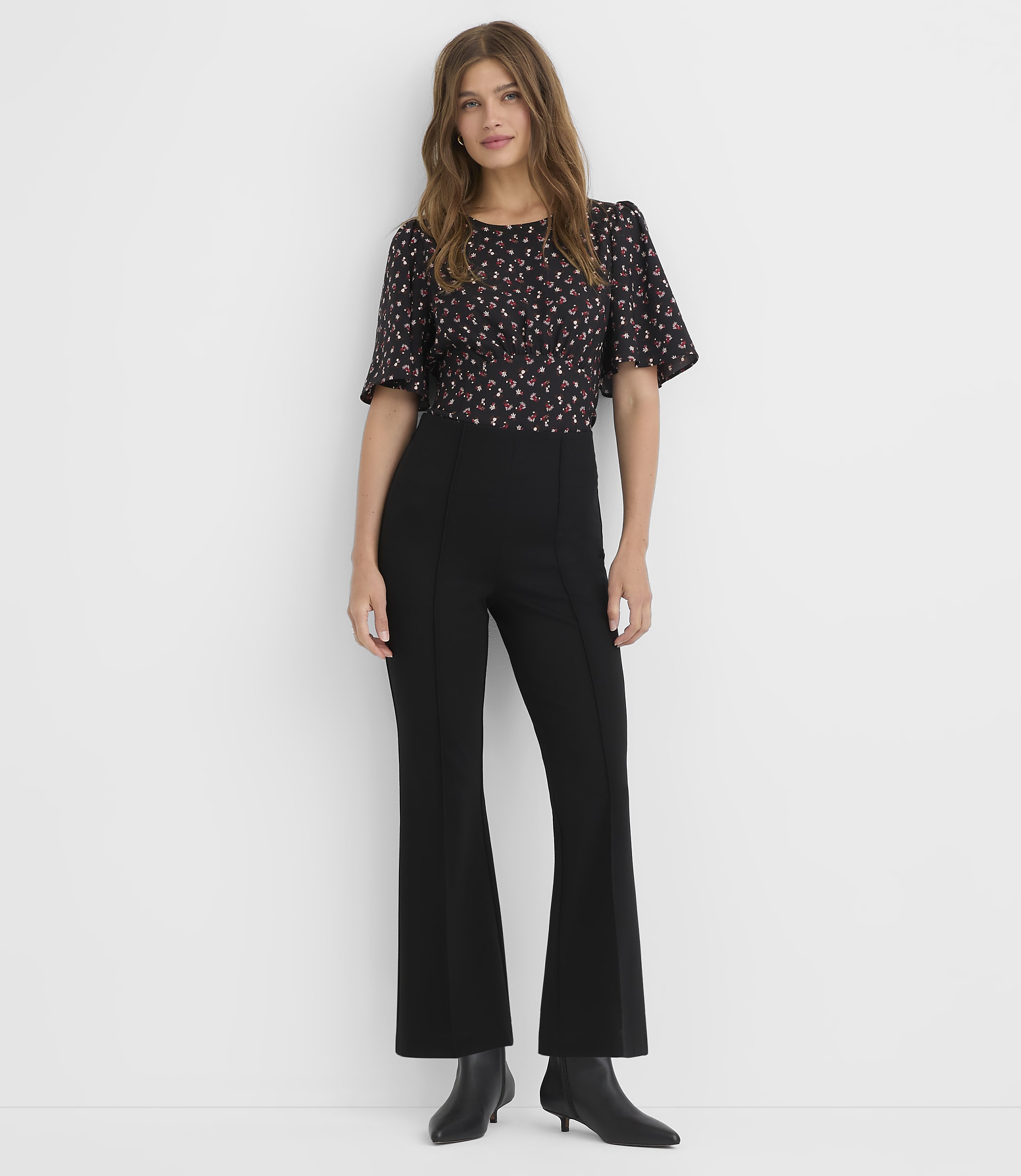 A woman in a floral top and black flared pants stands against a plain white background.